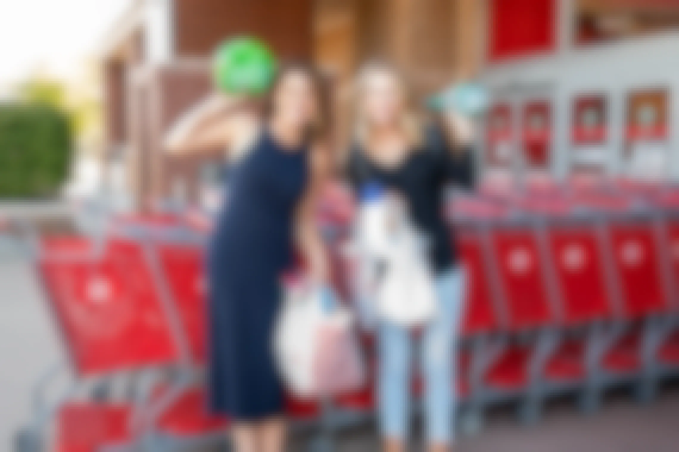 Two women standing outside of a Target storefront in front of a line of Target shopping carts. Both women are holding Target shopping bags and holding up a product that was purchased there.