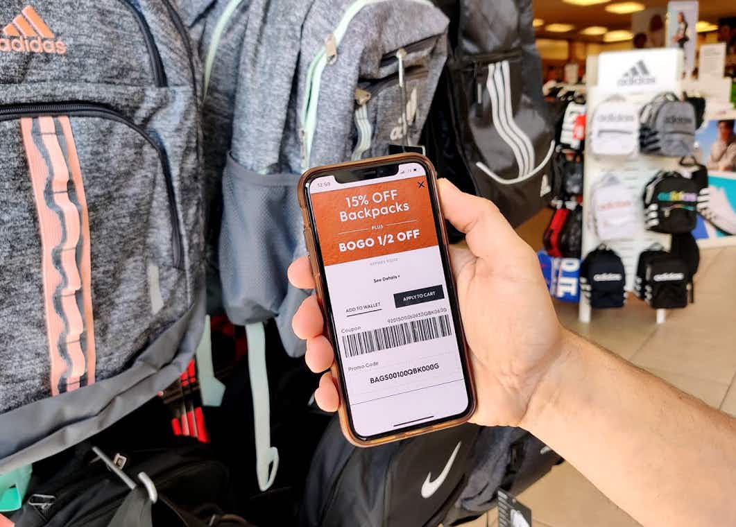 A phone held up in front of a rack of backpacks that reads 15% off backpacks and Buy one Get one