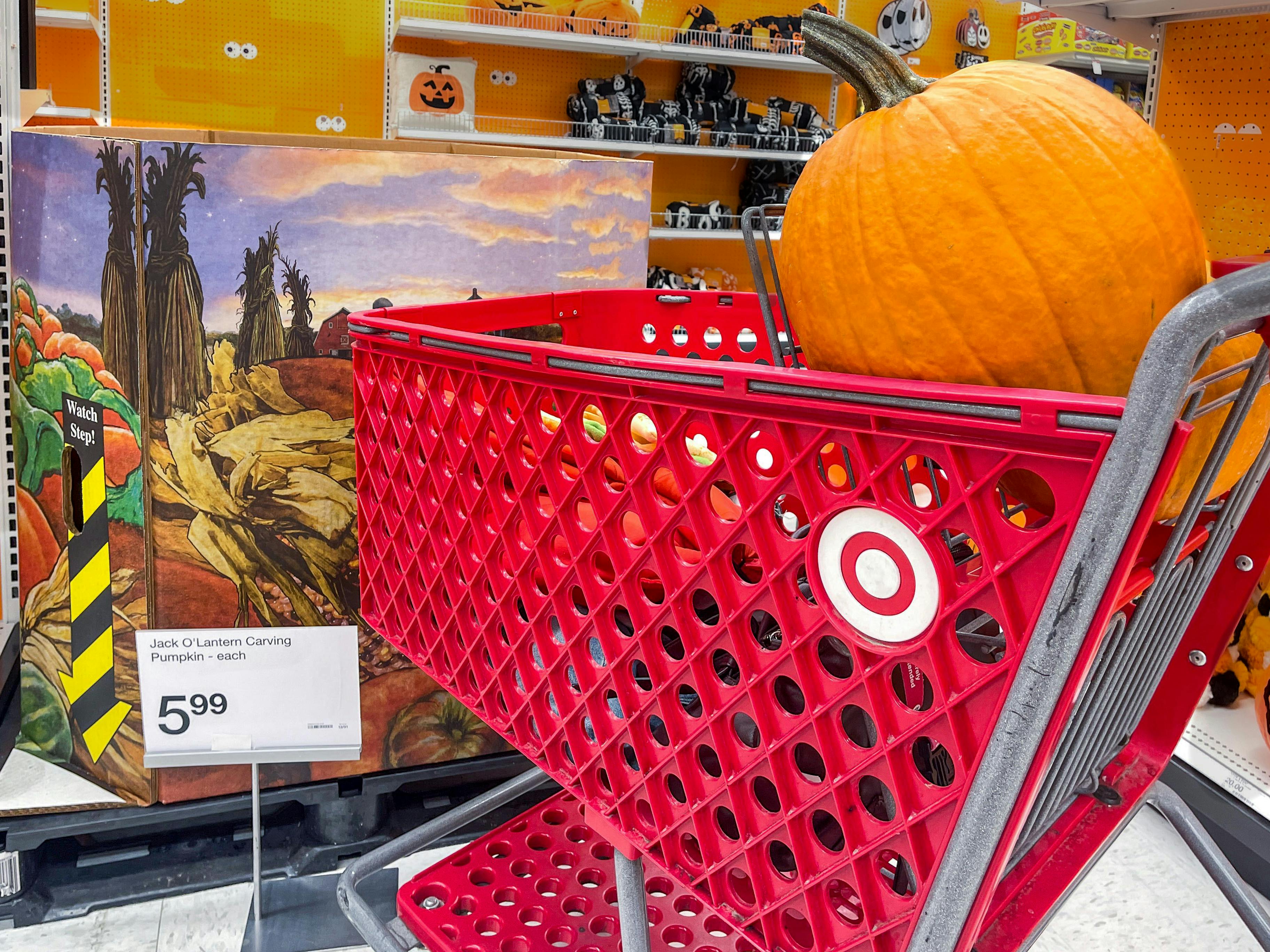 A pumpkin in a Target shopping cart parked next to a large box of more pumpkins and a sign that shows the price to be $5.99