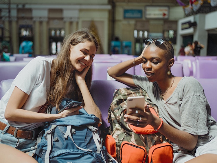 two friends sitting in airport with luggage and looking at their phones