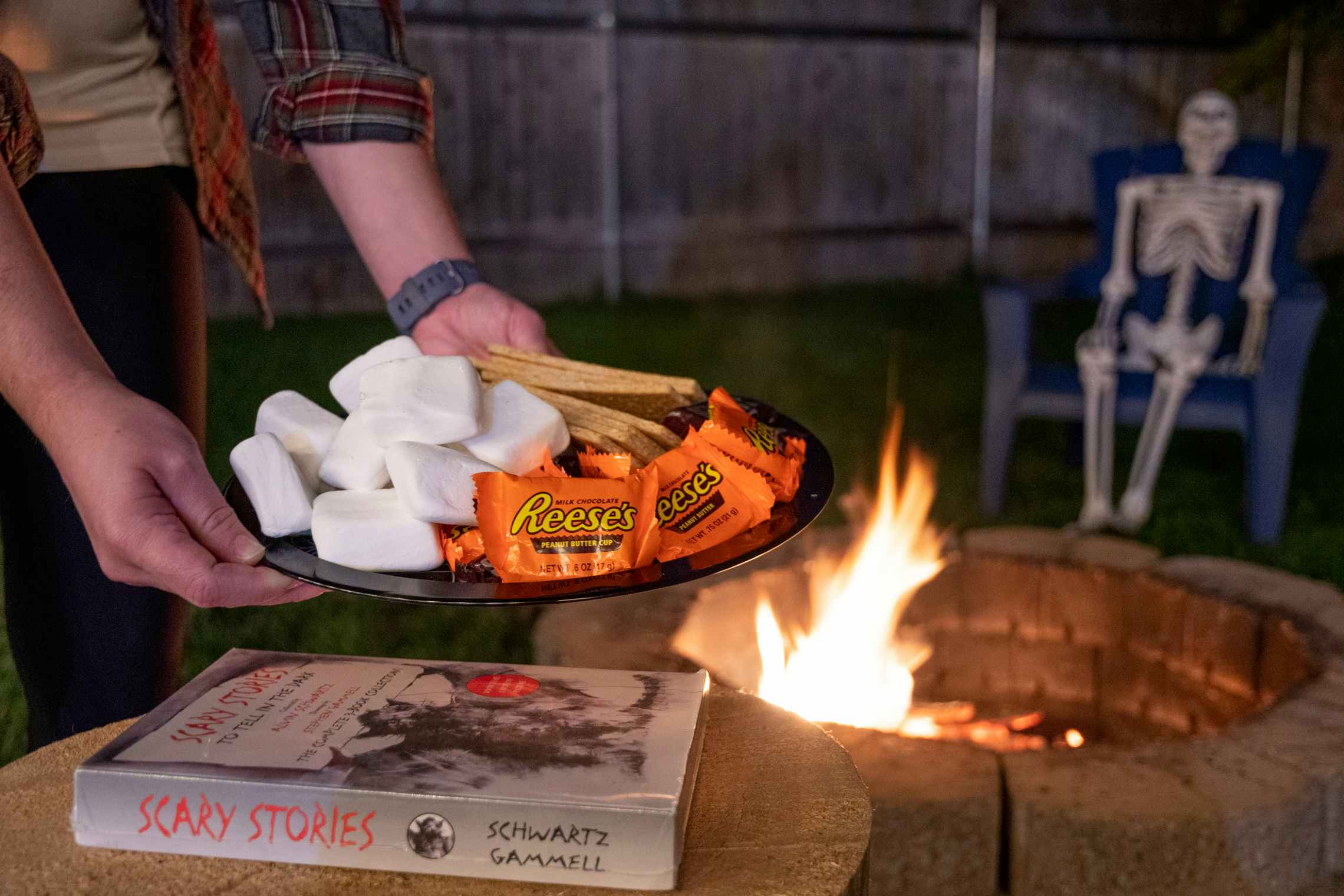 A person holding a plate of graham crackers, chocolate Reese's peanut butter cups and, marshmallows next to a fire pit and Scary Stories books, with a plastic skeleton on a chair in the background.