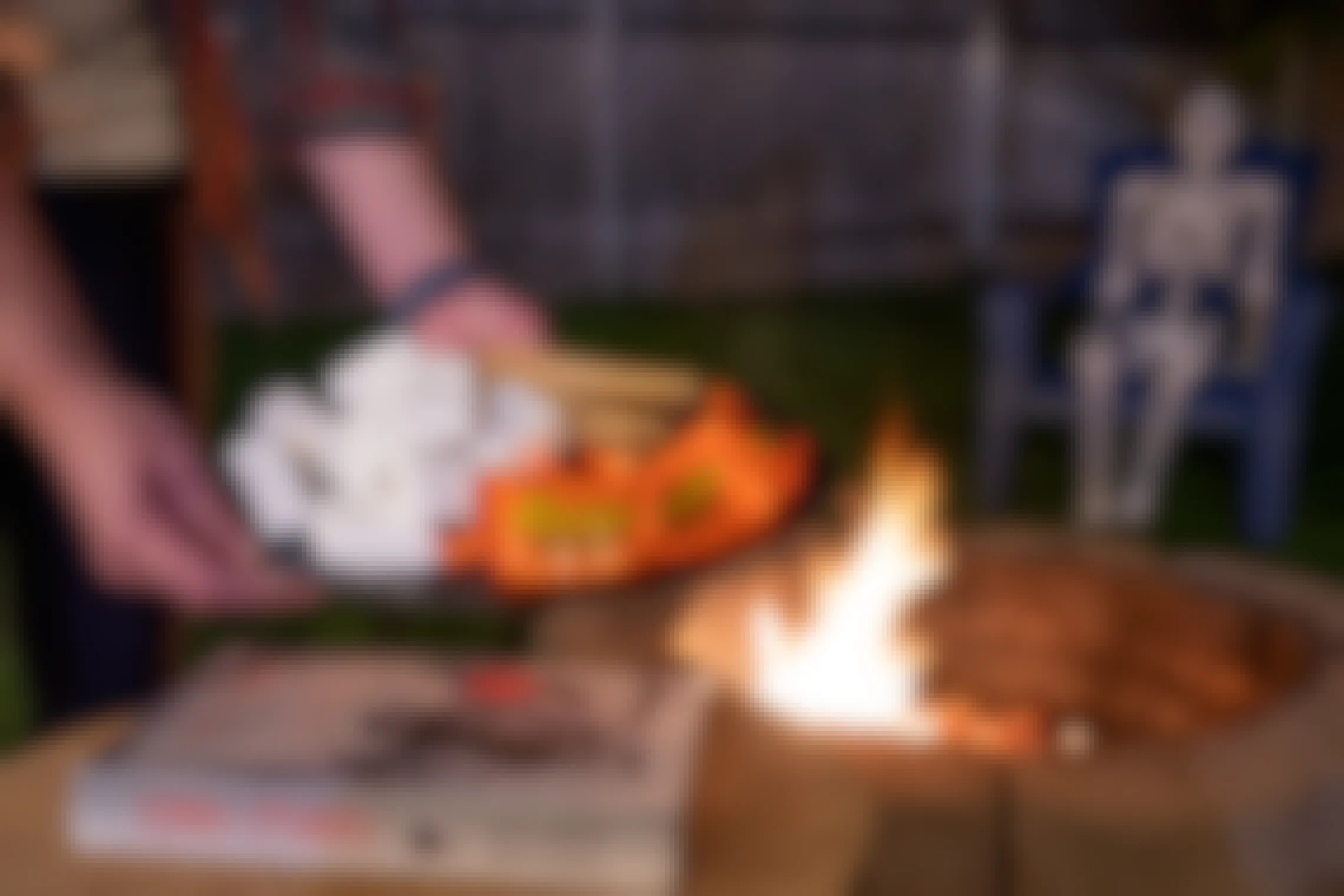 A person holding a plate of graham crackers, chocolate Reese's peanut butter cups, and marshmallows next to a fire pit and Scary Stories books, with a plastic skeleton sitting on a chair in the background..