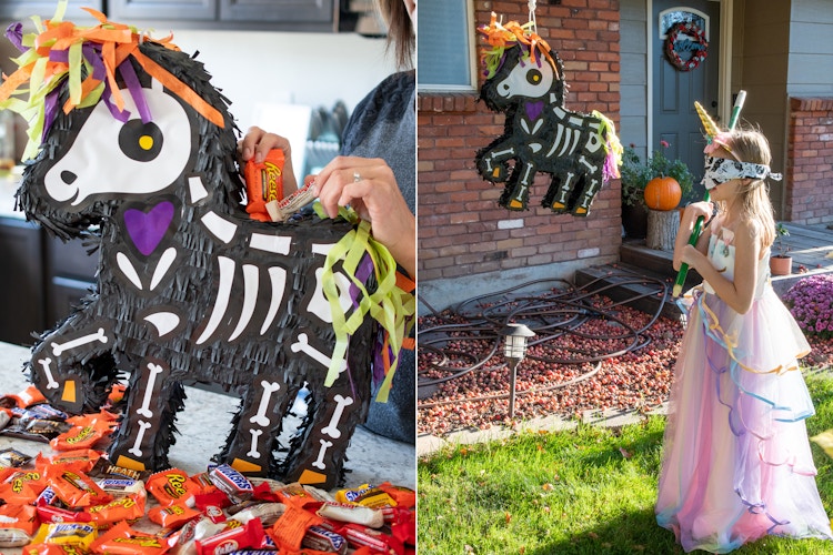 A woman filling a Halloween skeleton unicorn piñata with candy and a little girl wearing a blind fold and a halloween costume, holding a broom stick, ready to hit the piñata.