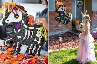 A woman filling a Halloween skeleton unicorn piñata with candy and a little girl wearing a blind fold and a halloween costume, holding a broom stick, ready to hit the piñata.