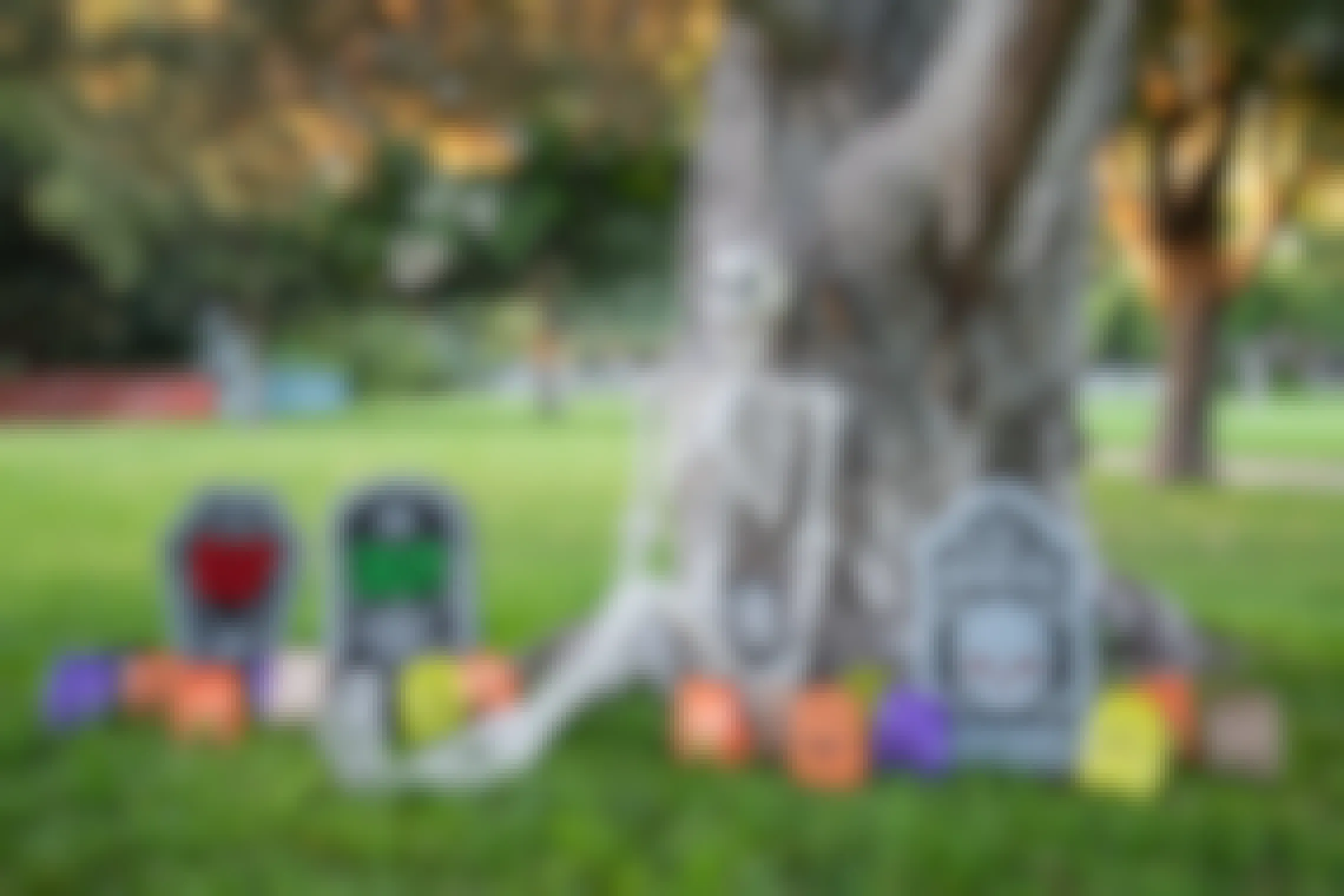 A skeleton learning against a tree among three decoration tombstones and Halloween trick-or-treat paper bags.