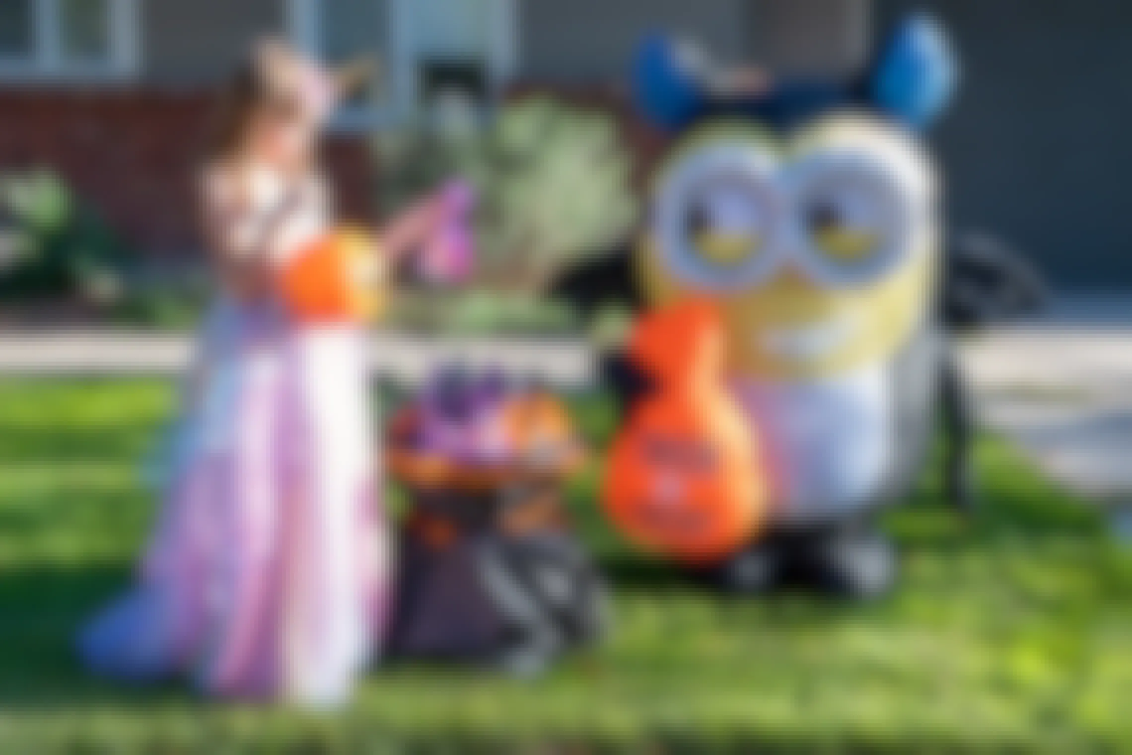 a little girl wearing a halloween costume, picking up a plastic treat bag from a table next to an inflatable minion lawn decoration.