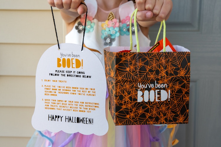 A little girl holding a sign shaped like ghost and a treat bag that say "you've been booed!