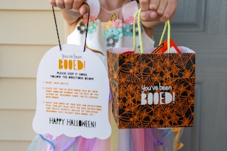 A little girl holding a sign shaped like ghost and a treat bag that say "you've been booed!