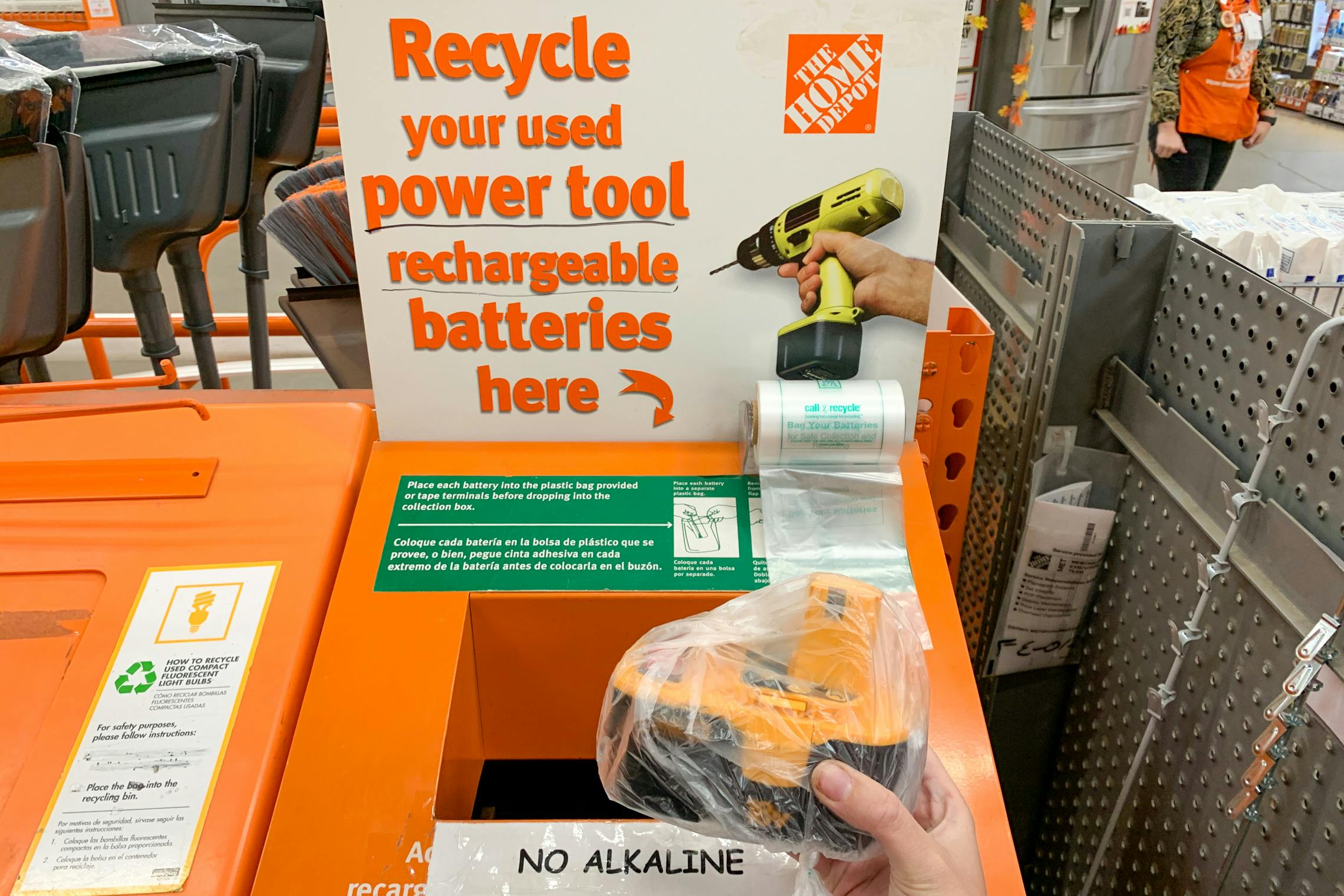 A person holding a rechargeable power tool battery in a plastic bag, in front of the recycle bin at home depot.