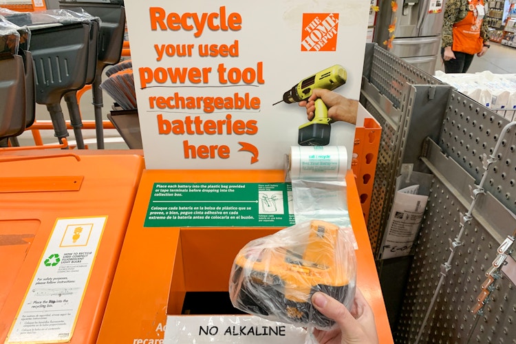 A person holding a rechargeable power tool battery in a plastic bag, in front of the recycle bin at home depot.
