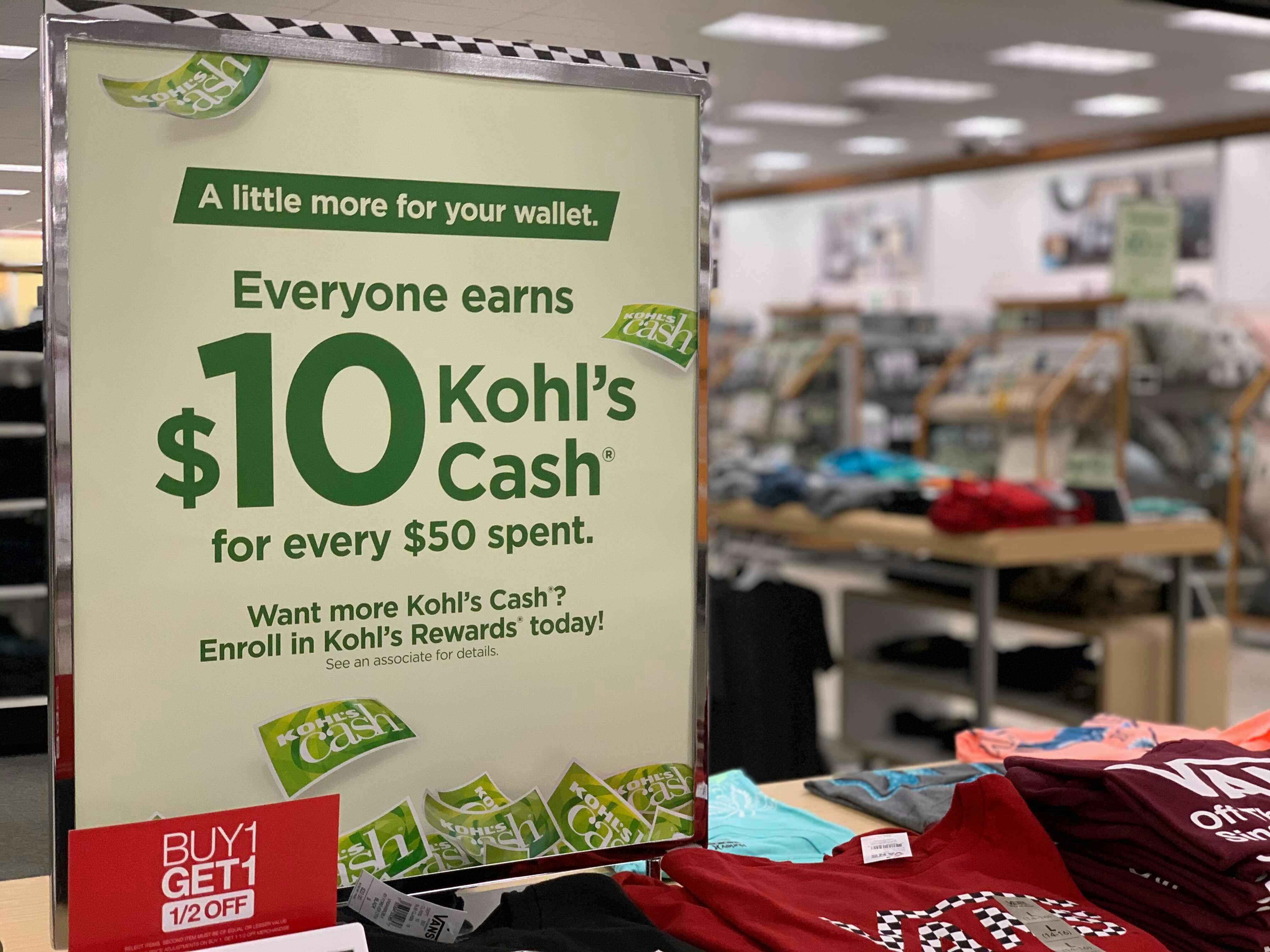 Kohls in store sign that reads Everyone earns $10 Kohls Cash for every $50 spent near a buy 1 get one ½ off sign on a tee shirt display stand