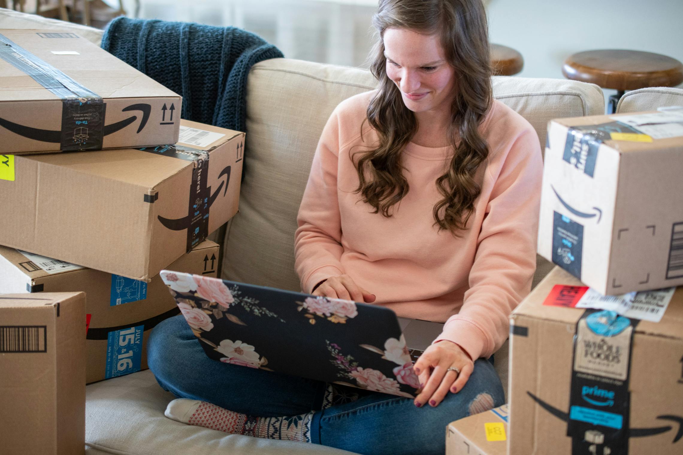 A woman sitting on a couch with a laptop computer on her lap, surrounded by stacks of Amazon delivery boxes.