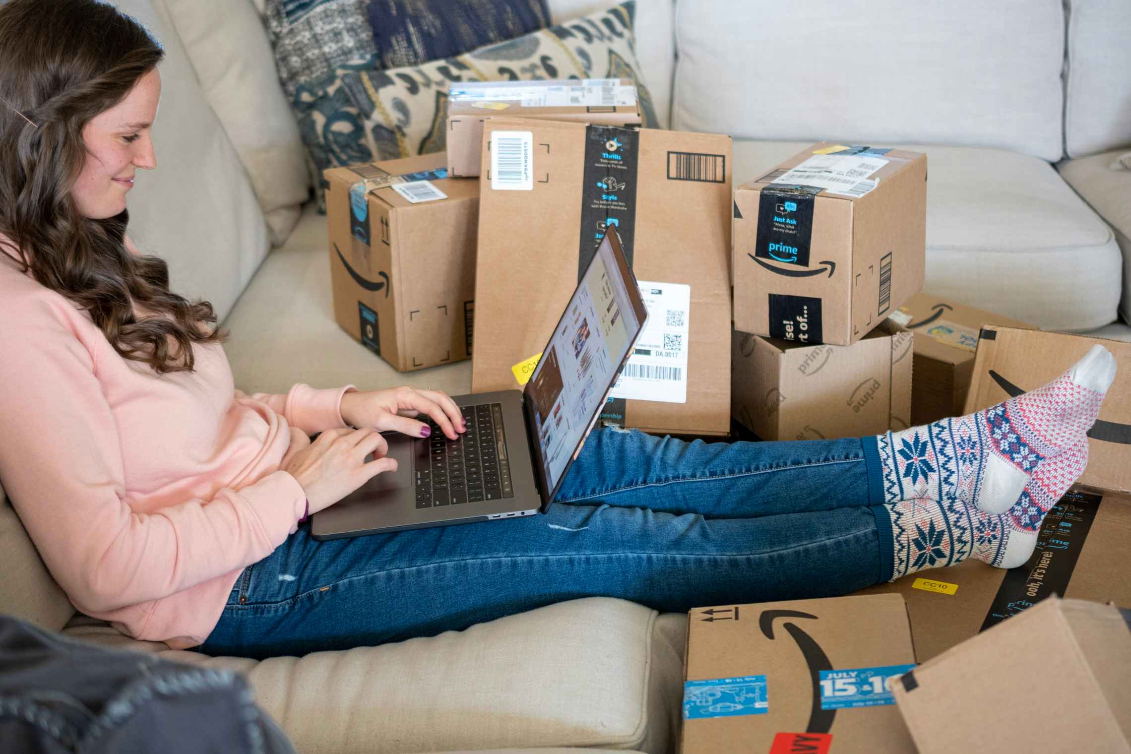 A woman sitting on a couch with a laptop computer on her lap, surrounded by Amazon delivery boxes.