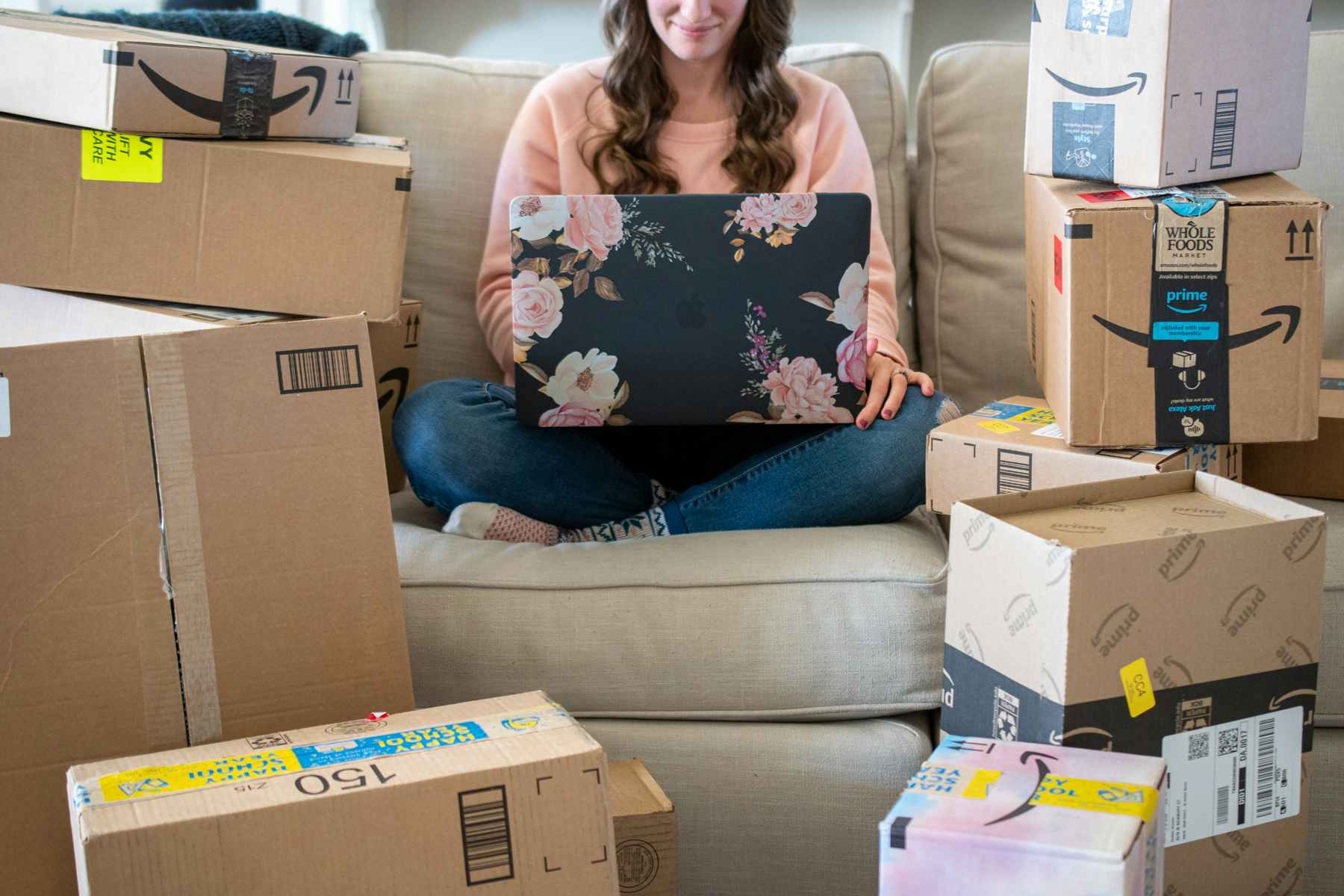 a woman sitting on her couch with her laptop surrounding by a dozen amazon prime packages of different sizes