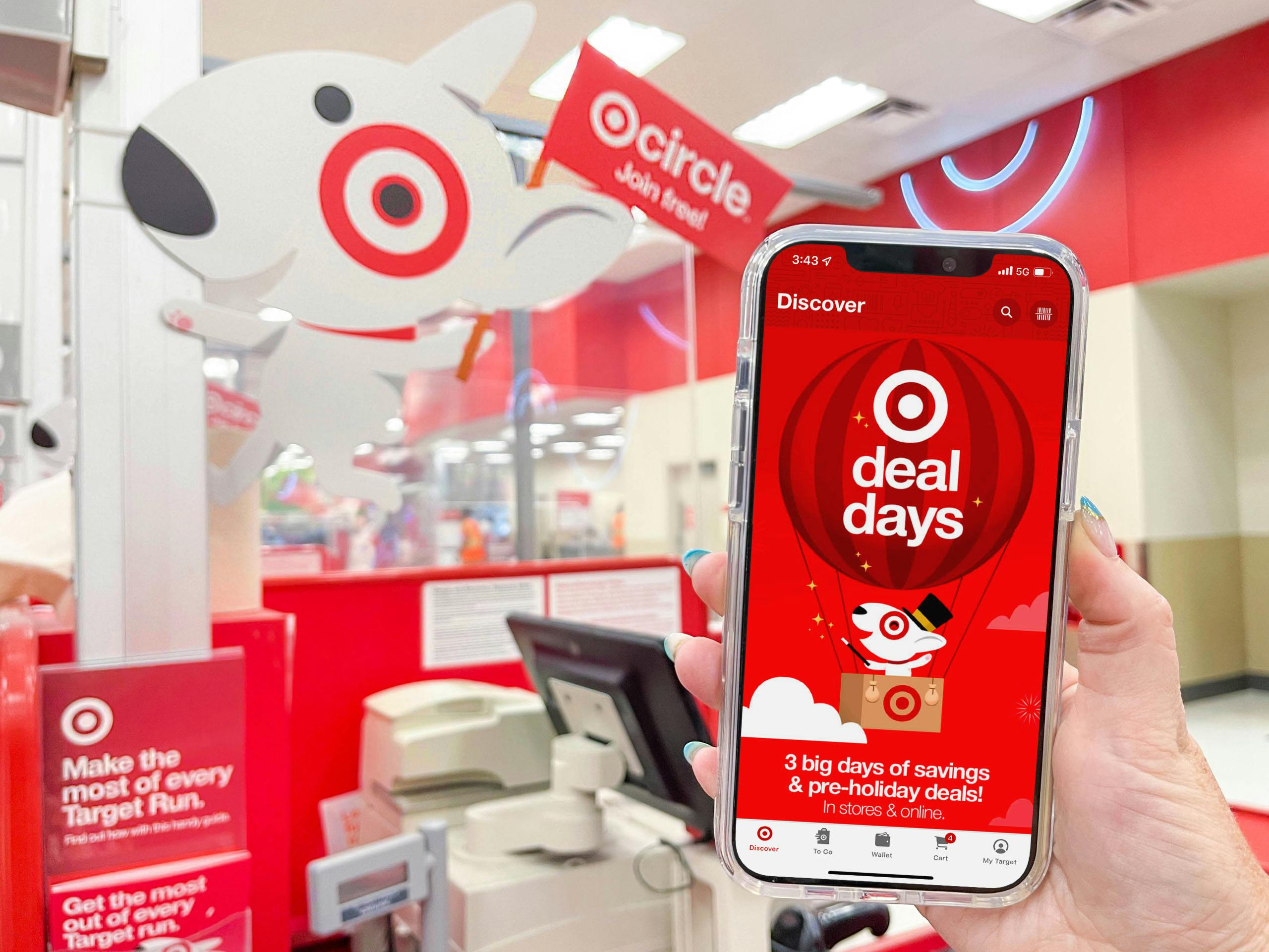 A person's hand holding up an iPhone displaying the Target app's Deal Days page in front of a Target checkout counter.