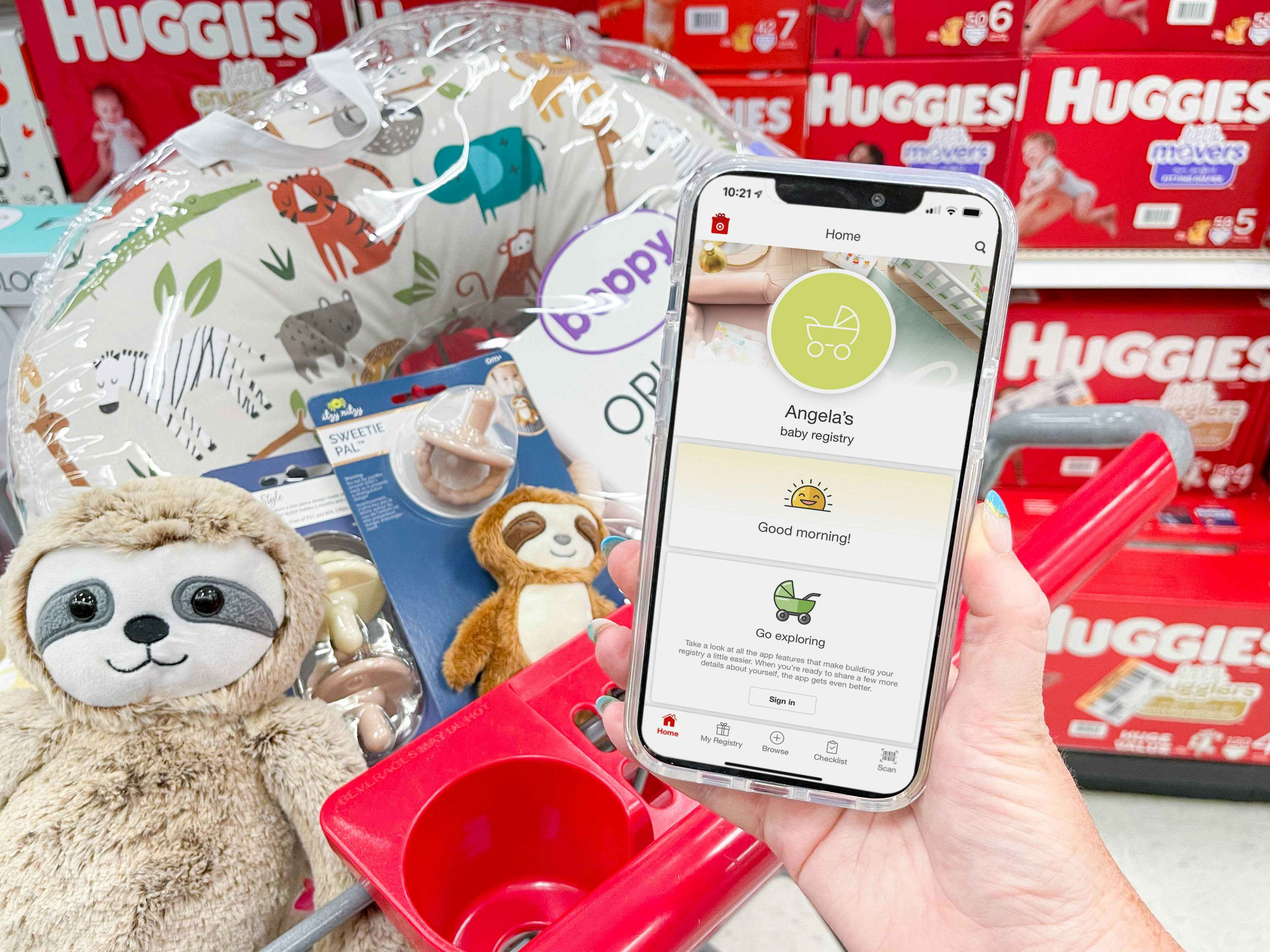 A person holding up their phone displaying a baby registry on the Target app in front of a Target shopping cart full of baby items.