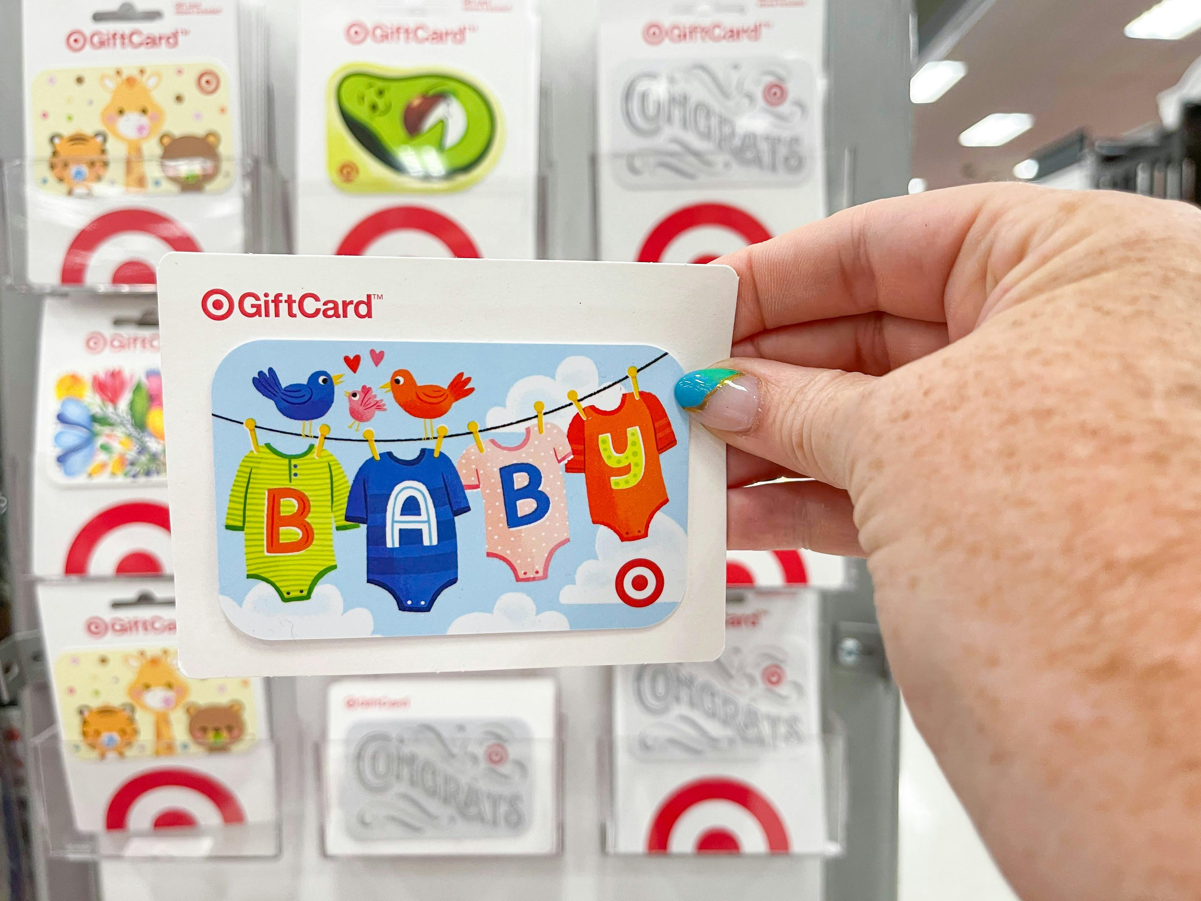 A person's hand holding up a baby-themed Target gift card in front of a display of gift cards.