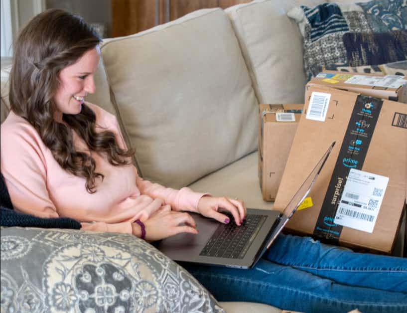 A woman sitting on a couch, looking at a computer with amazon packages around her.