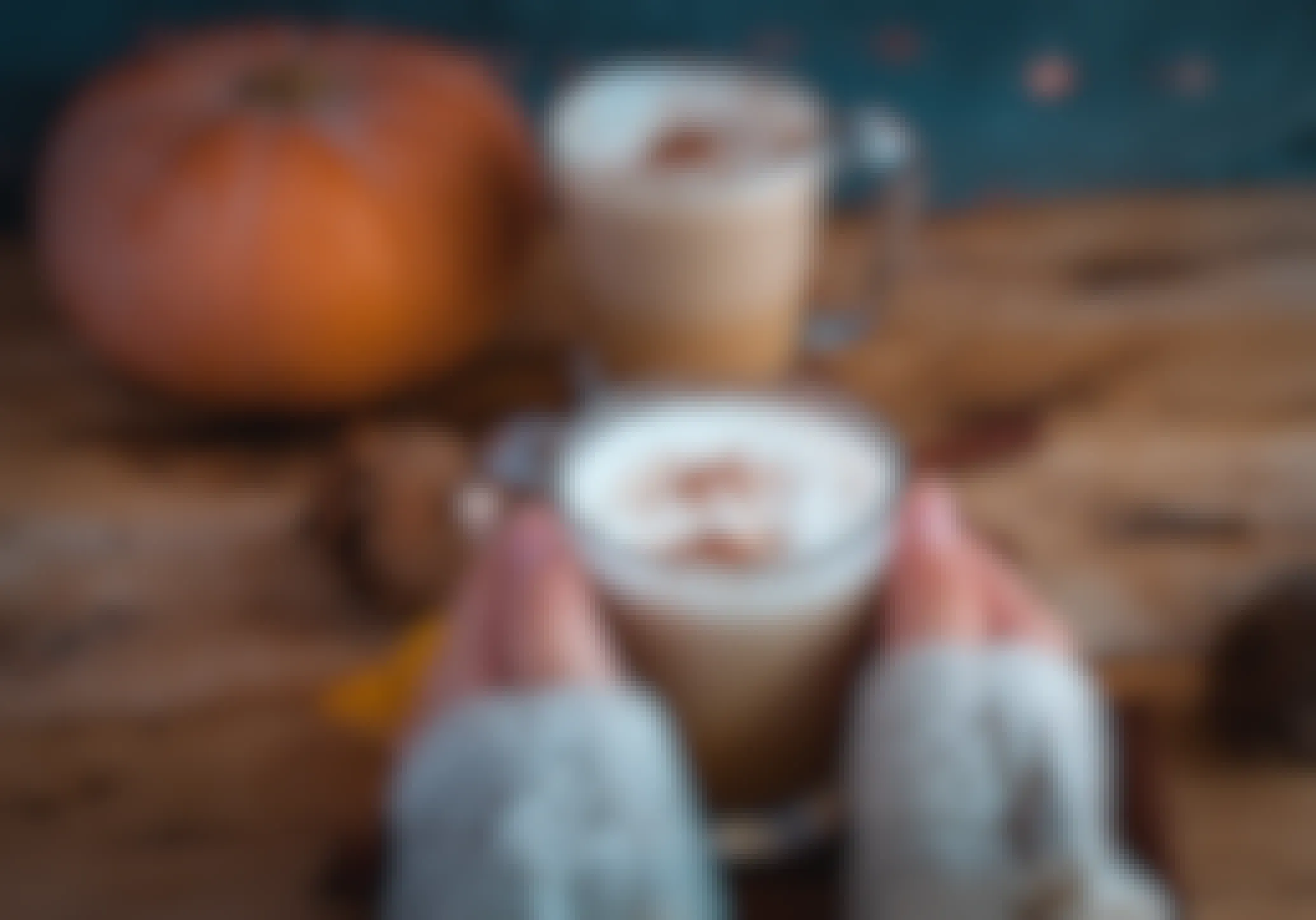 A person's hands holding a mug with a pumpkin spice drink next to a pumpkin on a table.