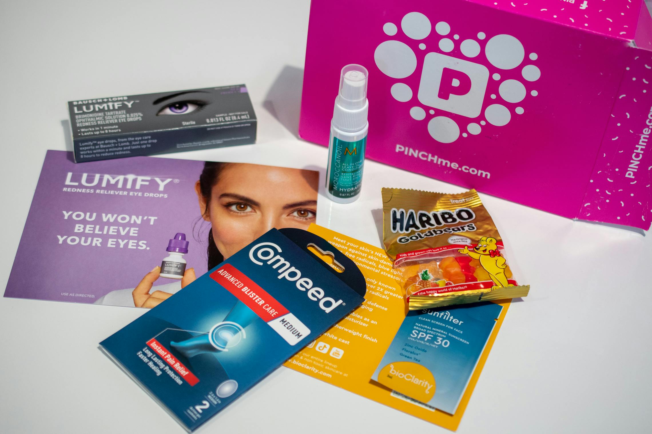 A pink box from PINCHme with free samples of products like Lumify, Compeed, and Haribo Gummy Bears scattered next to it.