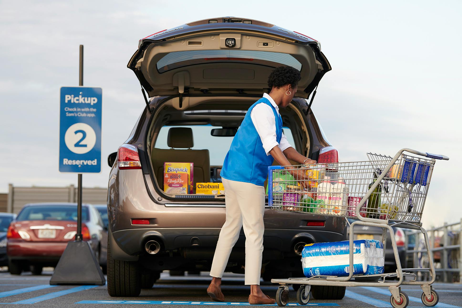 A Sam's Club employee packing groceries into the trunk of a vehicle that is parked in the Curbside pickup parking spot outside of Sam's Club.