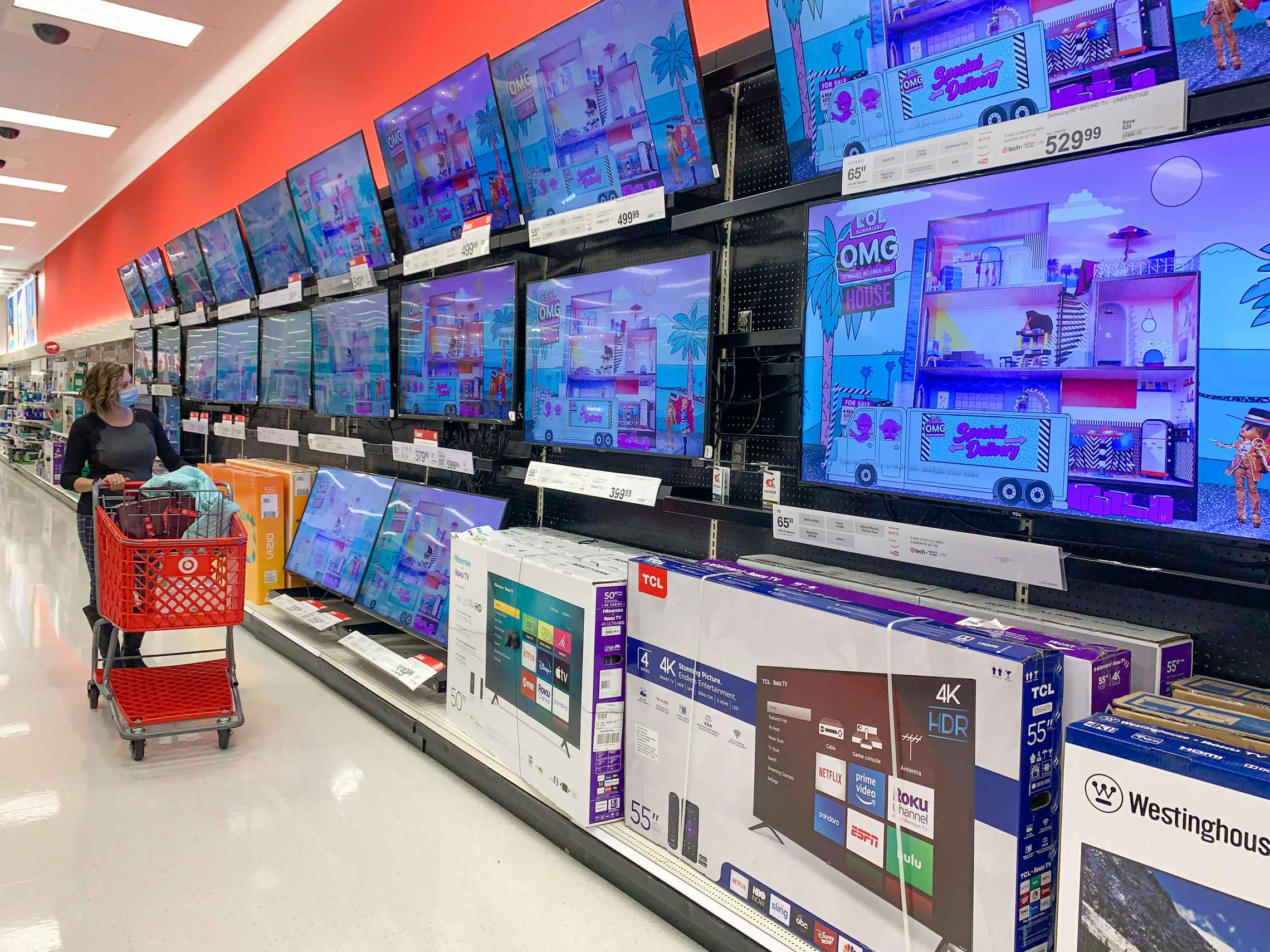 A woman pushing a Target shopping cart along a wall of display TVs in the electronics section at Target.