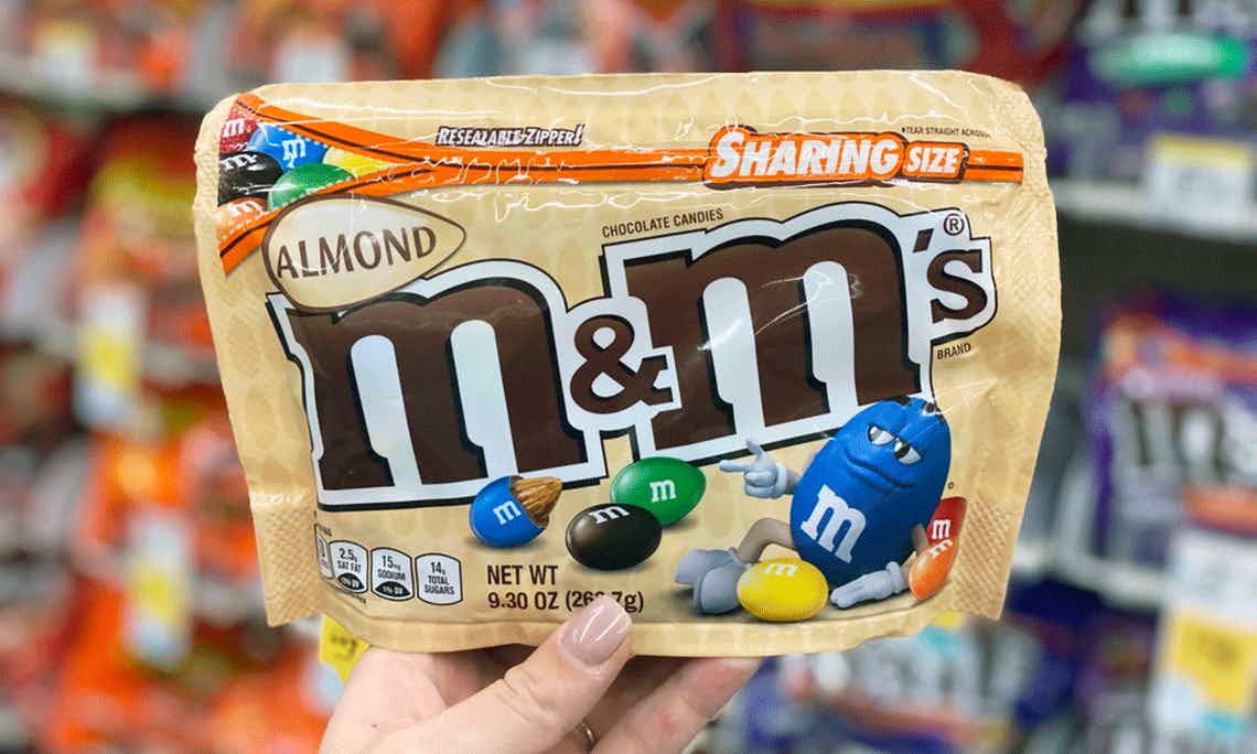 A person's hand holding up a bag of almond M&Ms in front of a candy shelf at Walgreens.