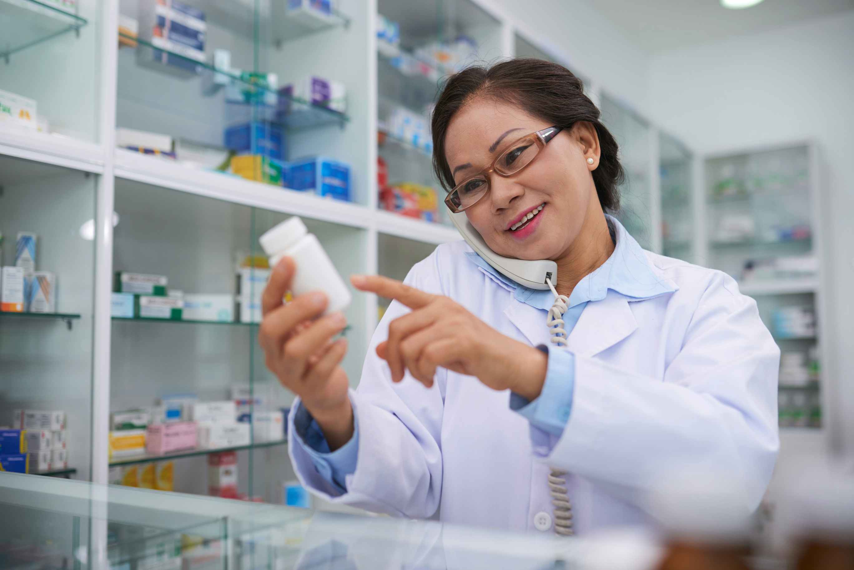 A pharmacist looking at a bottle of pills and talking on the phone.