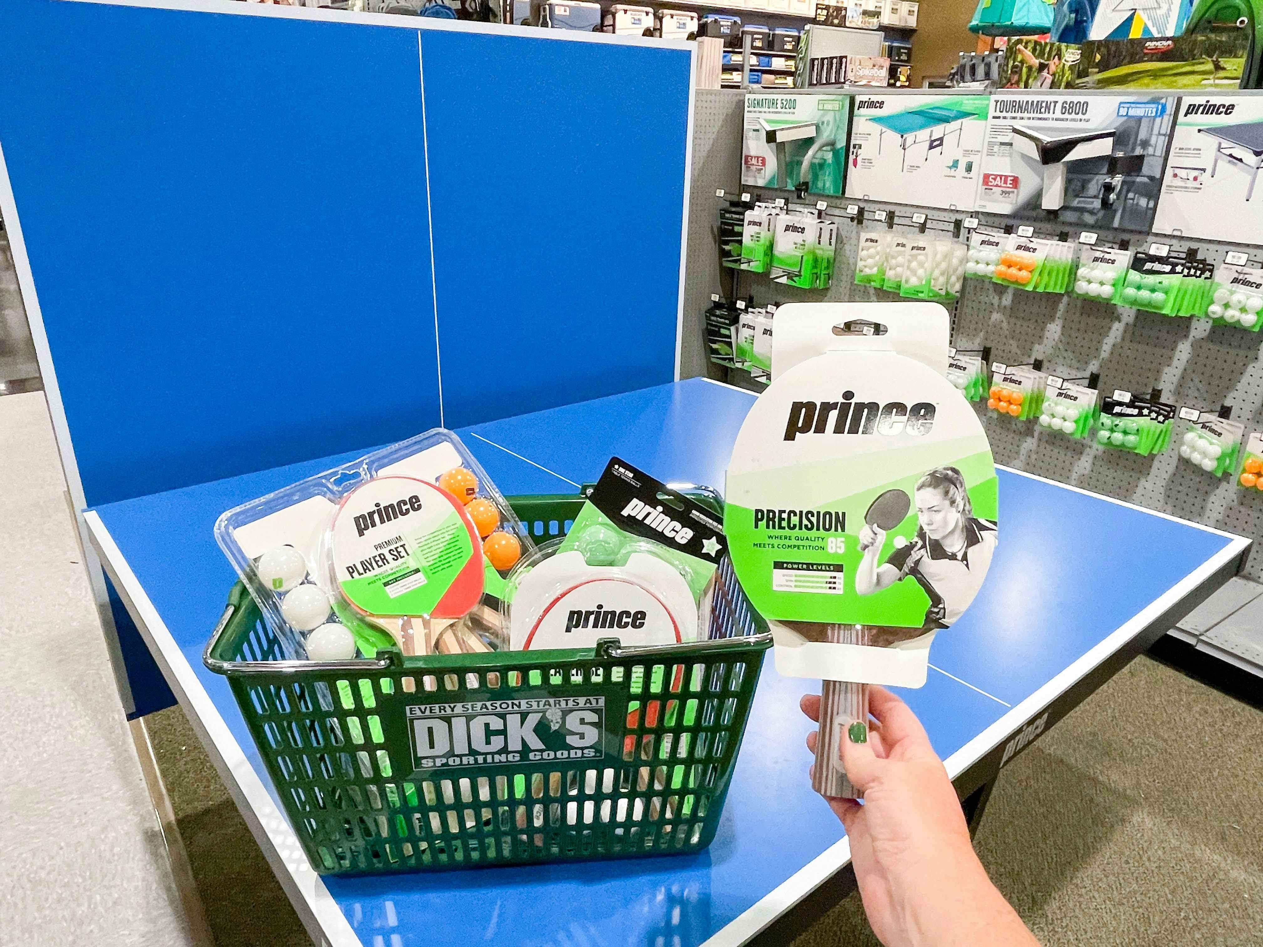 A person holding a packaged ping pong paddle in front of a Dick's Sporting Goods hand basket, filled with more ping pong paddles and balls, that sitting on a half-folded ping pong table.
