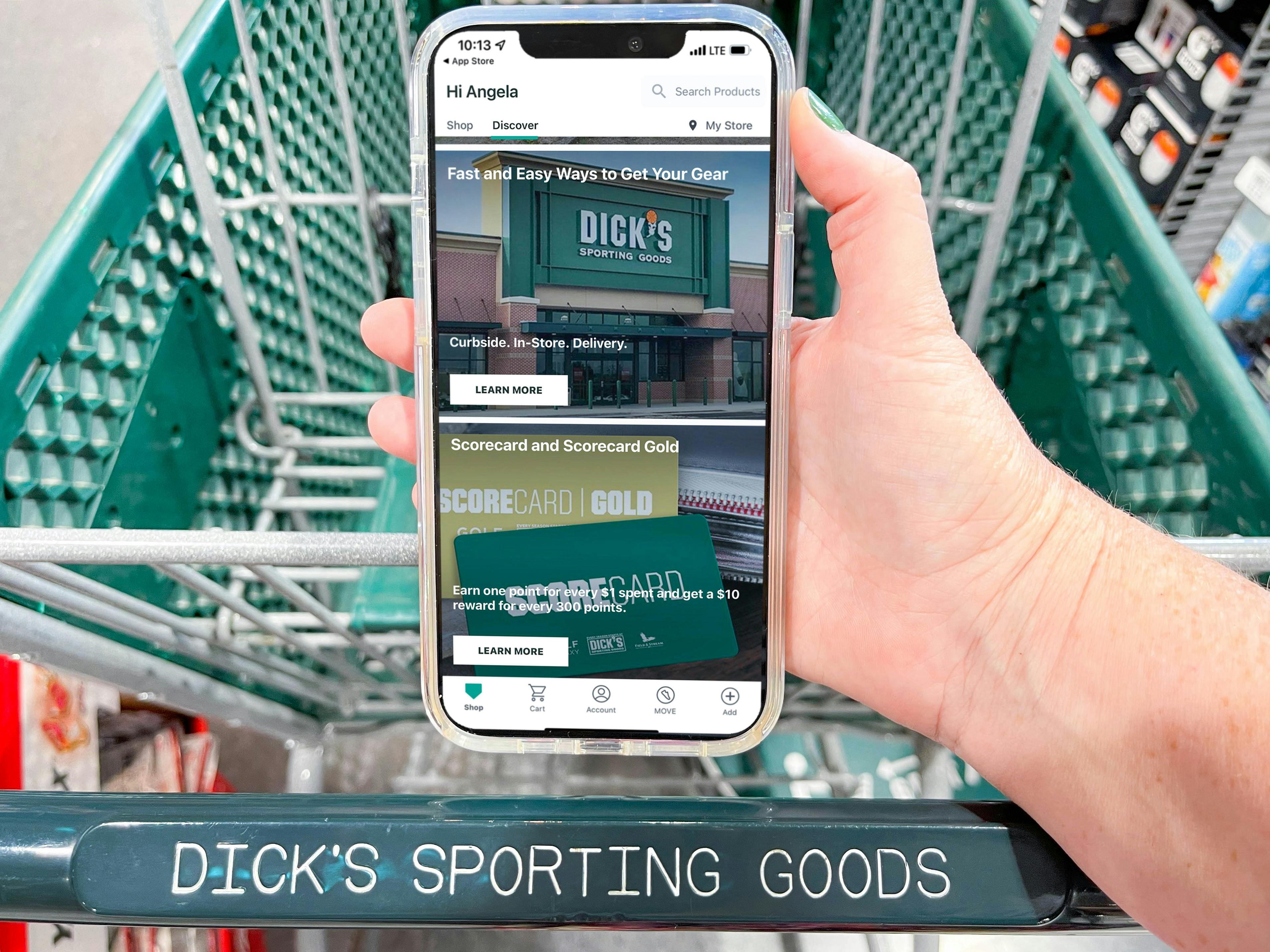 A person's hand holding their phone displaying the Dick's Sporting Goods mobile app above a Dick's Sporting Goods shopping cart.