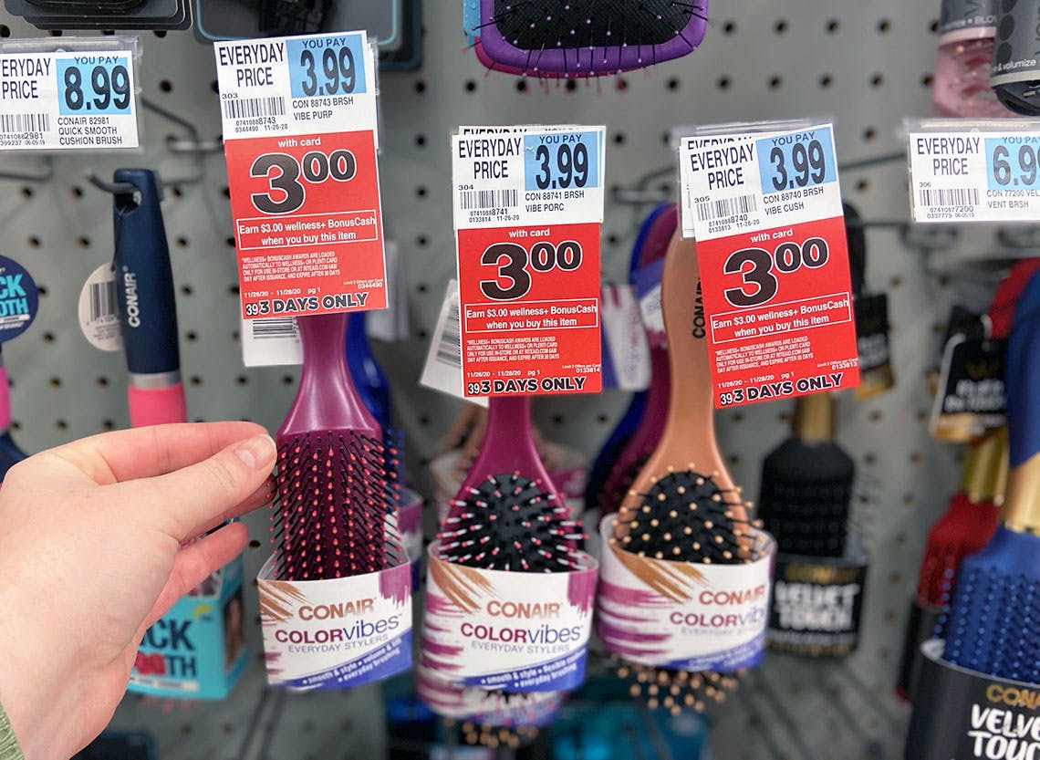 A person's hand reaching for a Conair colorvibes brush hanging on a display with BonusCash sale tags inside Rite Aid.