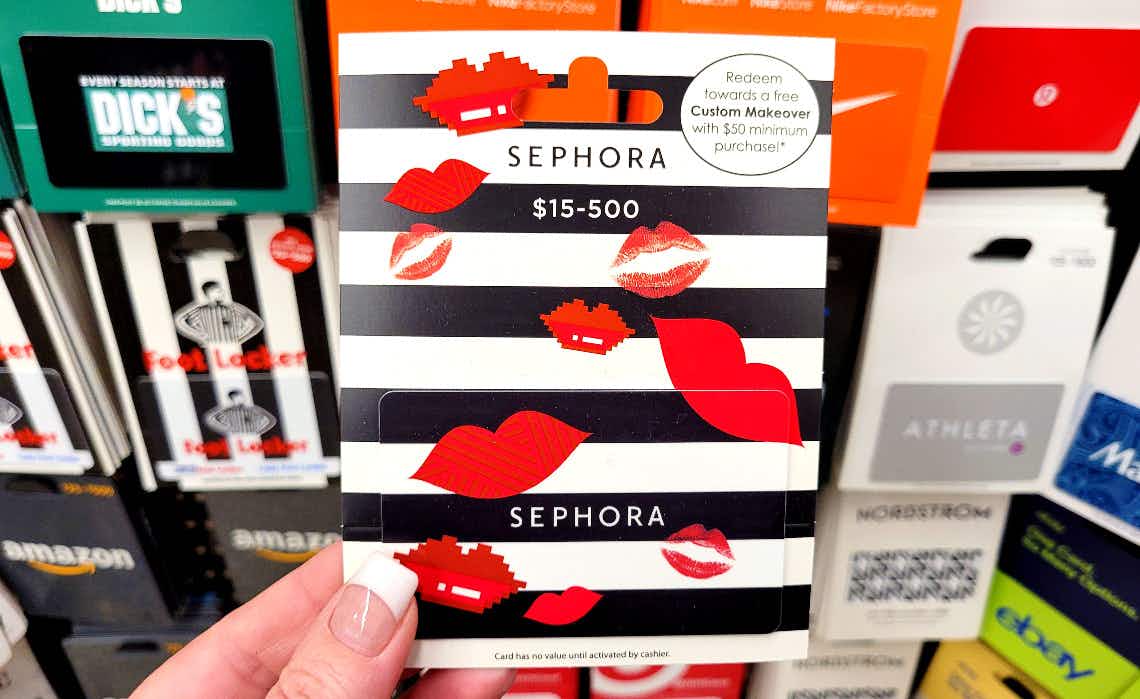A person's hand holding a Sephora gift card in front of a display of gift cards.