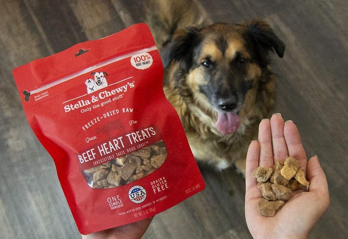 A person holding a bag of Stella & Chewy's dog treats in one hand and a handful of treats from the bag in the other hand, above a dog that is sitting and looking up toward the treats.