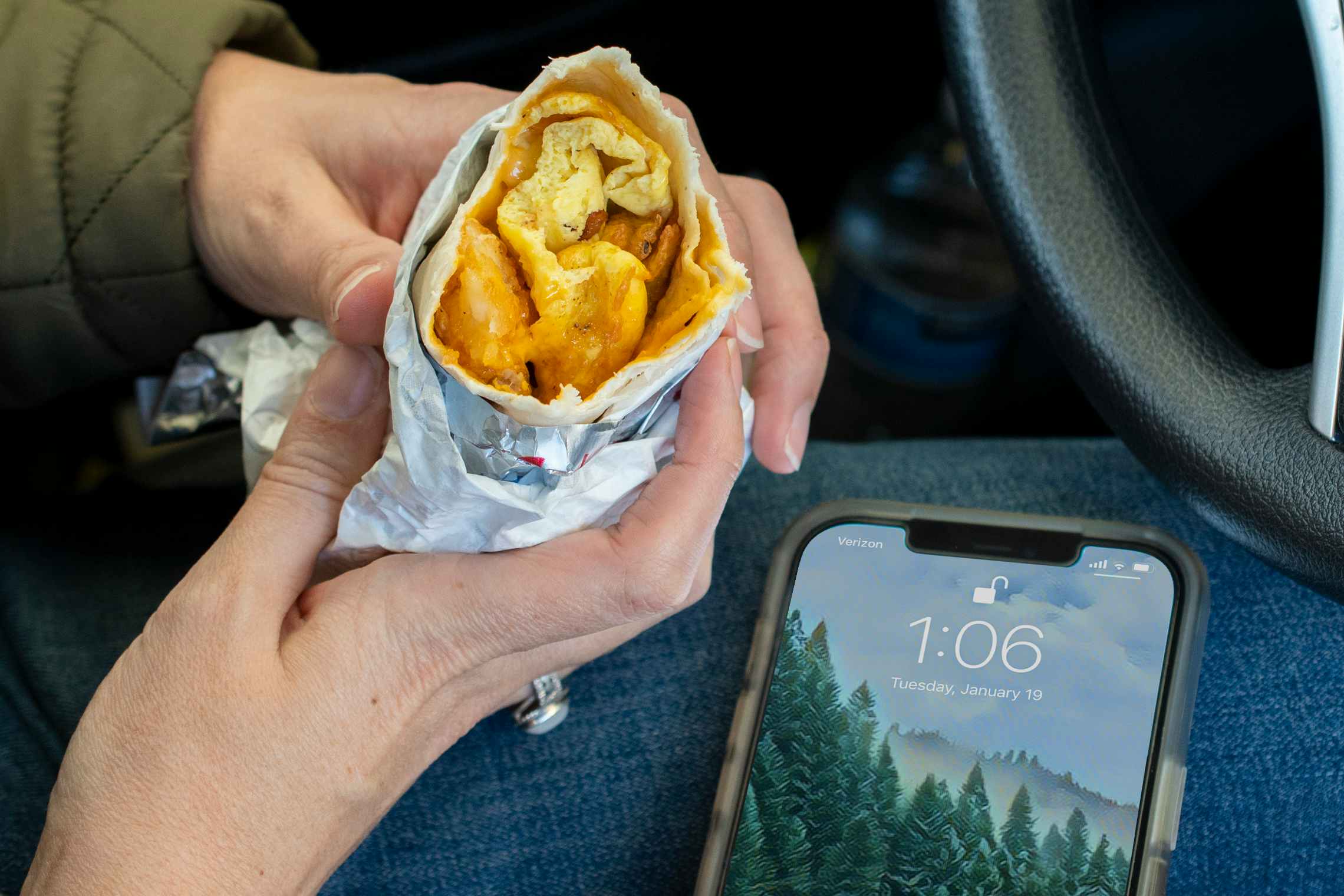 A person holding a Sonic breakfast burrito with a cell phone sitting on their lap displaying the time, 1:06 pm, on the screen.