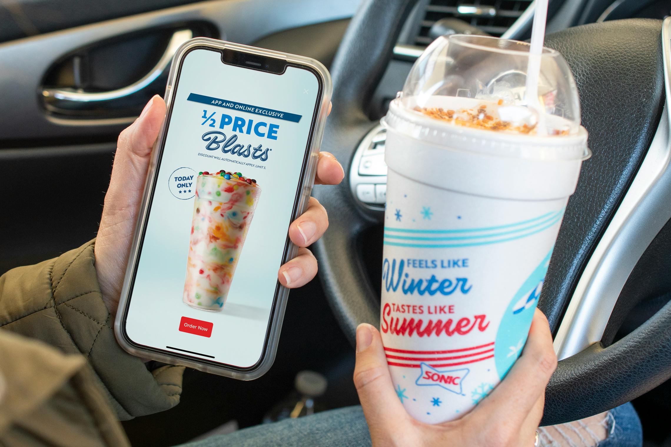 A person sitting in their car, holding a Sonic Blast cup next to their cell phone, displaying an advertisement in the Sonic app that reads, "1/2 price Blasts"