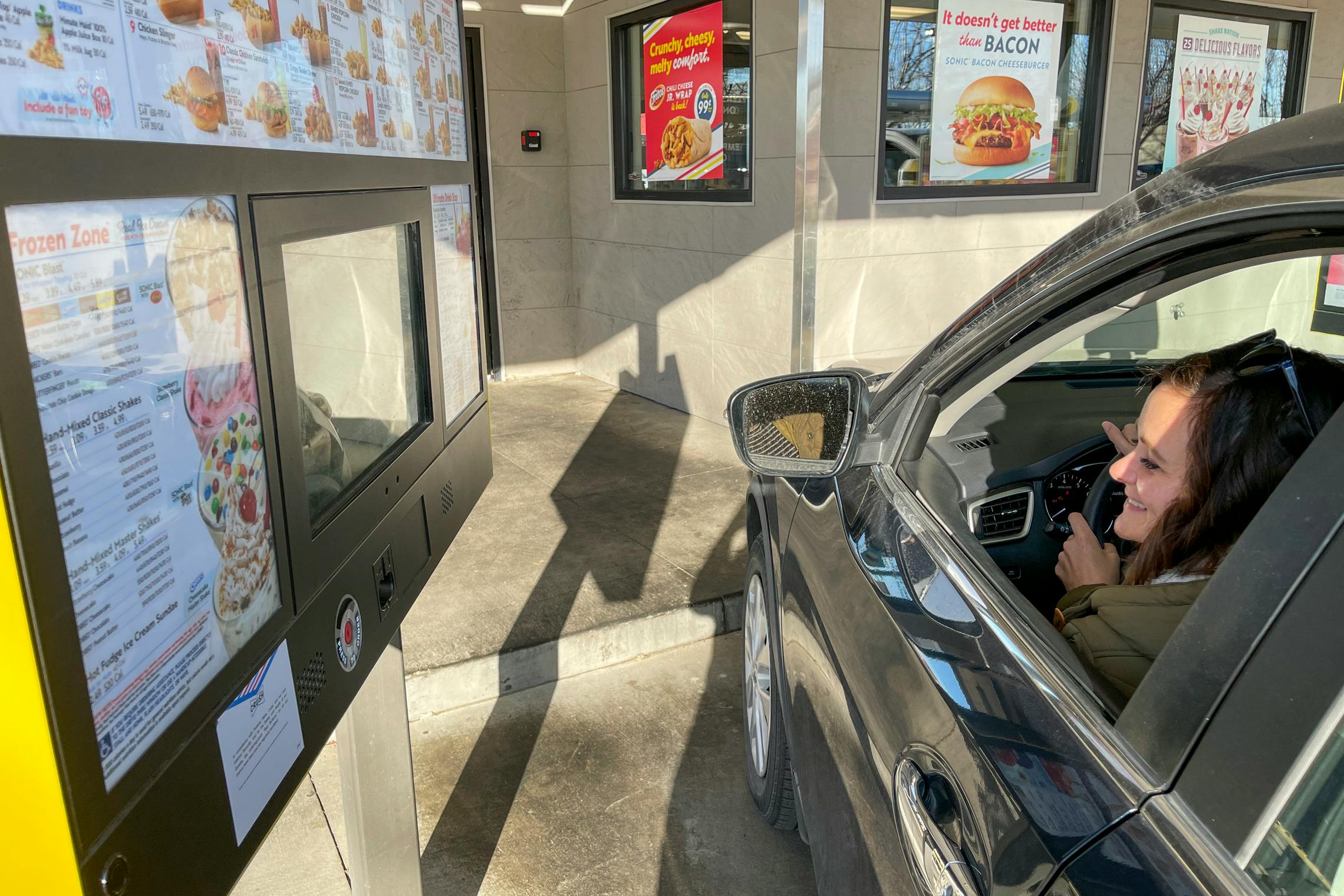A woman sitting in her vehicle with her window down, ordering from the a kiosk at a Sonic Drive-in.