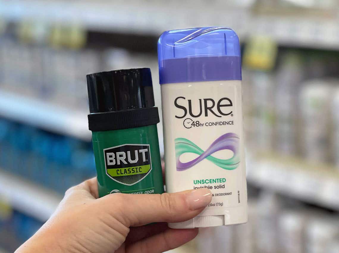 A person's hand holding up a stick of Sure deodorant and Brut Classic in front of a shelf in a CVS.