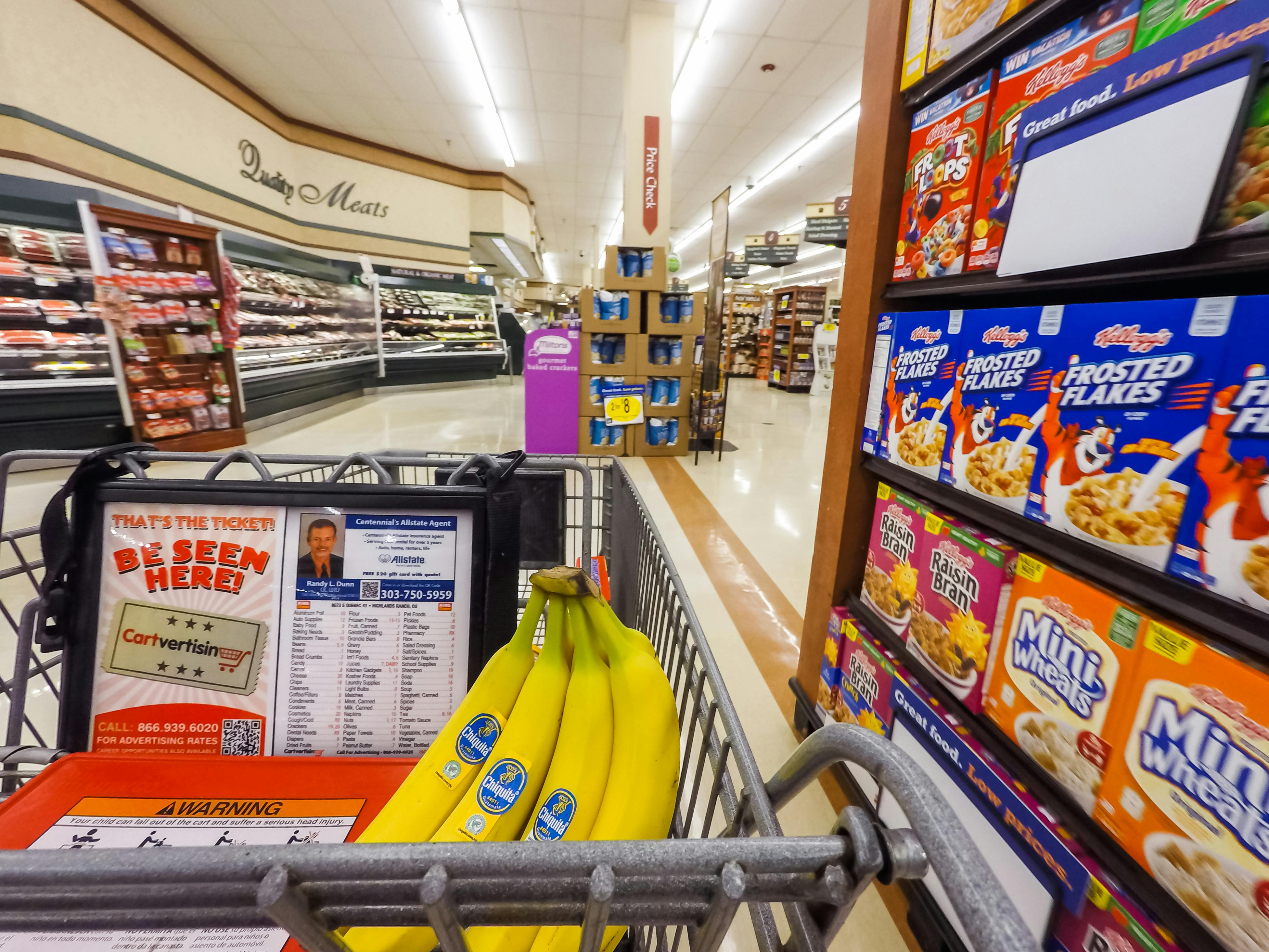 A shopping cart with bananas in the basket, next to a shelf filled with cereal.