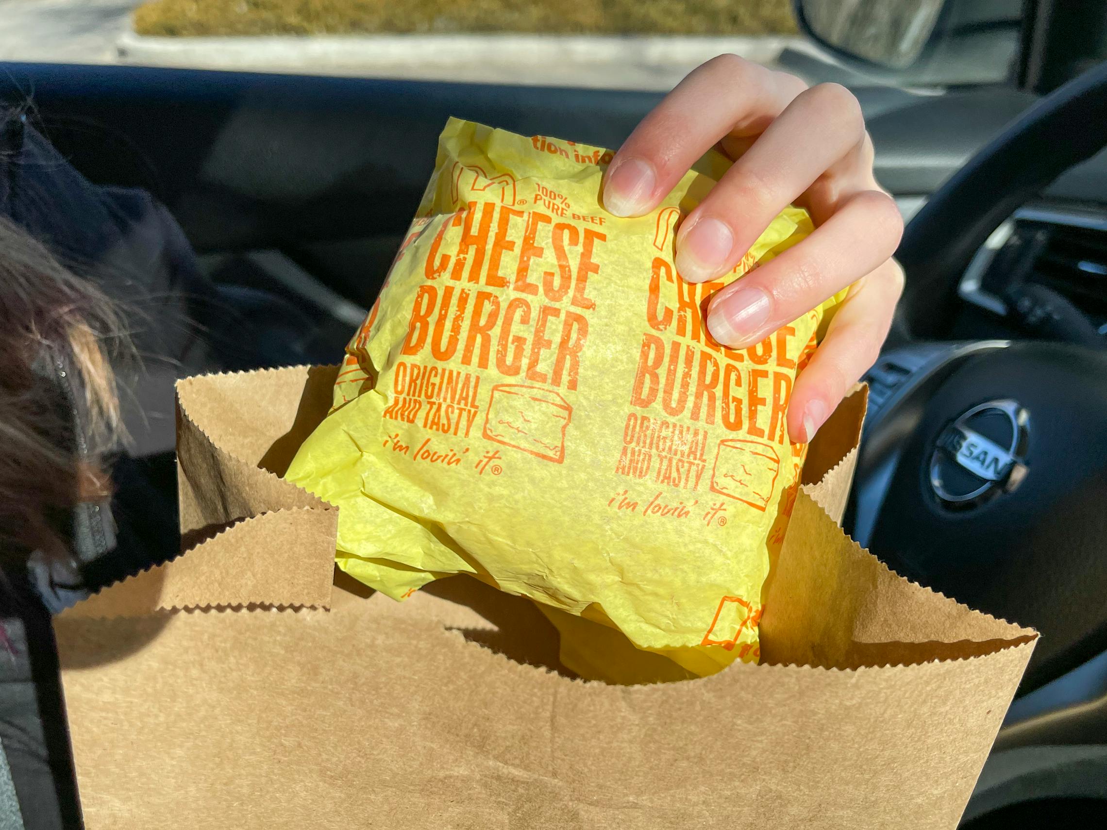 A person pulling a wrapped McDonald's cheeseburger out of a McDonald's bag while sitting in their car.