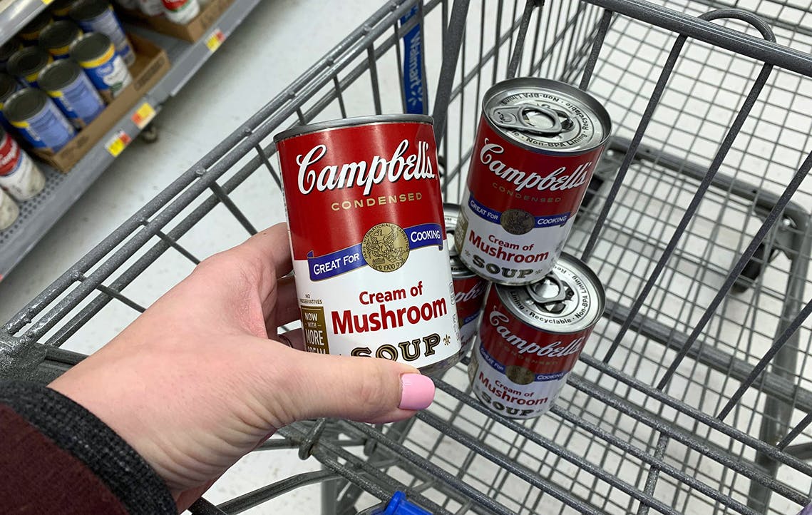 A person's hand placing a can of Campbell's cream of mushroom soup into a Walmart shopping cart basket with other cans of Campbell's soup.