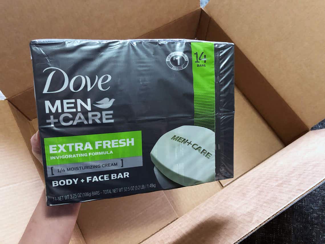 A hand holding a pack of Dove Men+Care soap above an open Amazon delivery box.