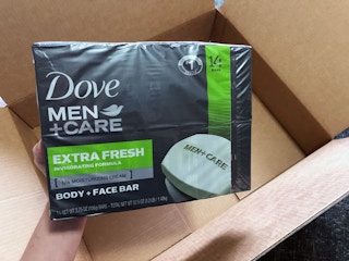 A hand holding a pack of Dove Men+Care soap above an open Amazon delivery box.