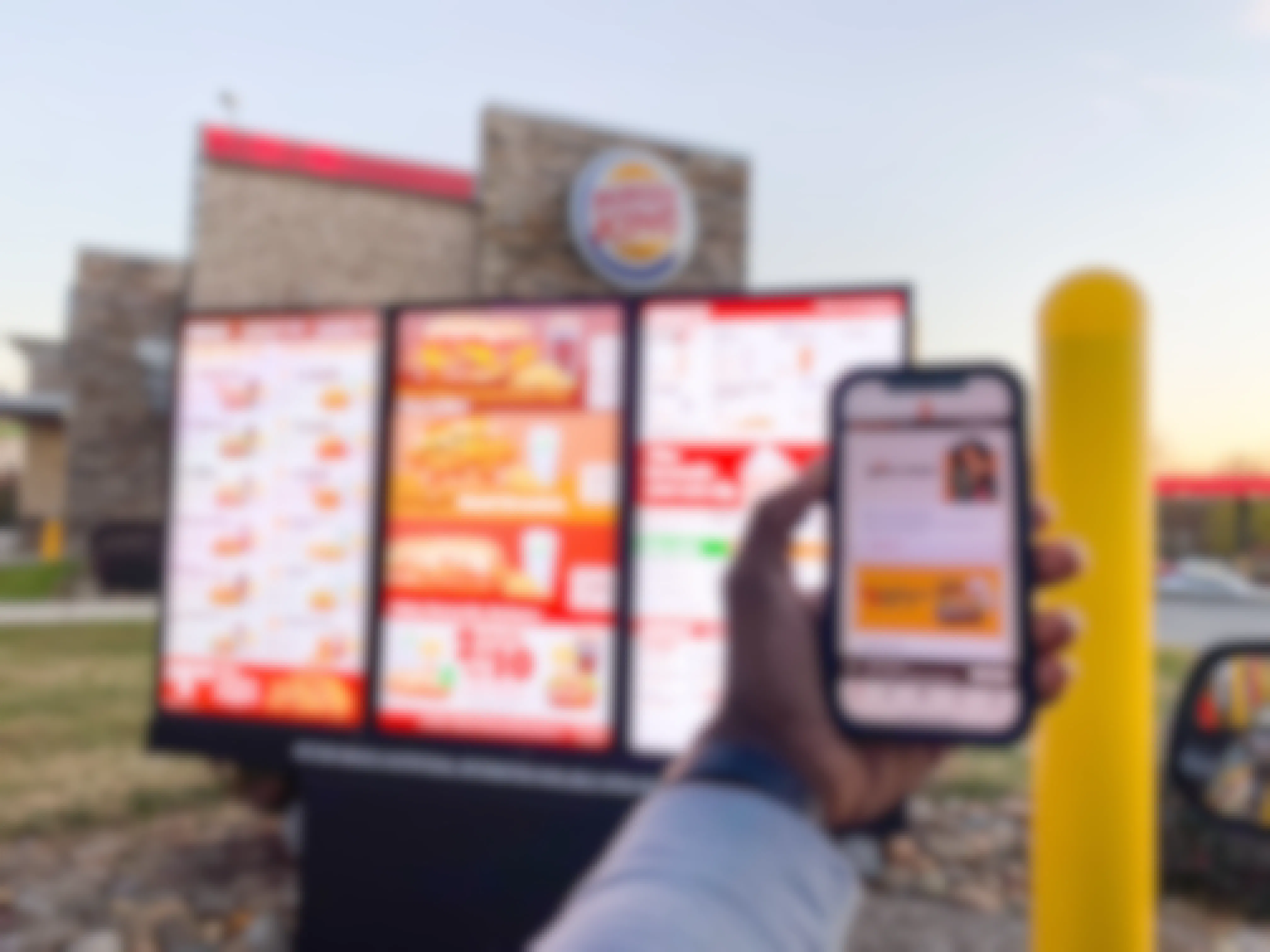A person's hand holding up their phone displaying the Burger King Royal Perks app next to the menu in the Burger King drive thru line.