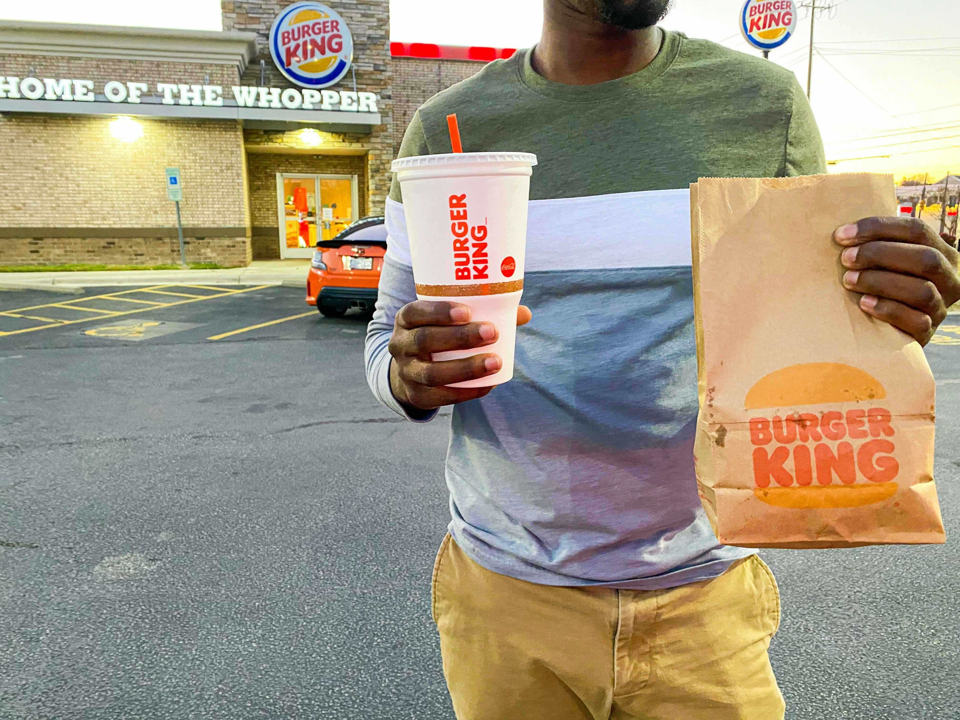 A person walking with a Burger King drink and takeout bag in front of a Burger King restaurant.