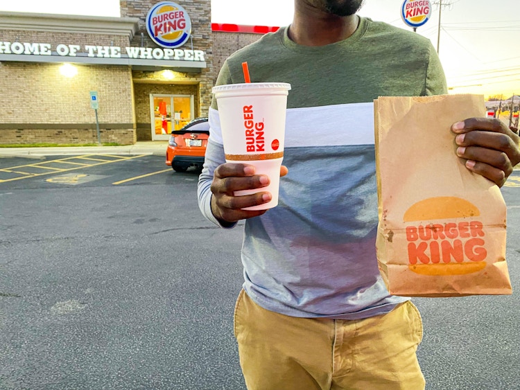 A person walking with a Burger King drink and takeout bag in front of a Burger King restaurant.