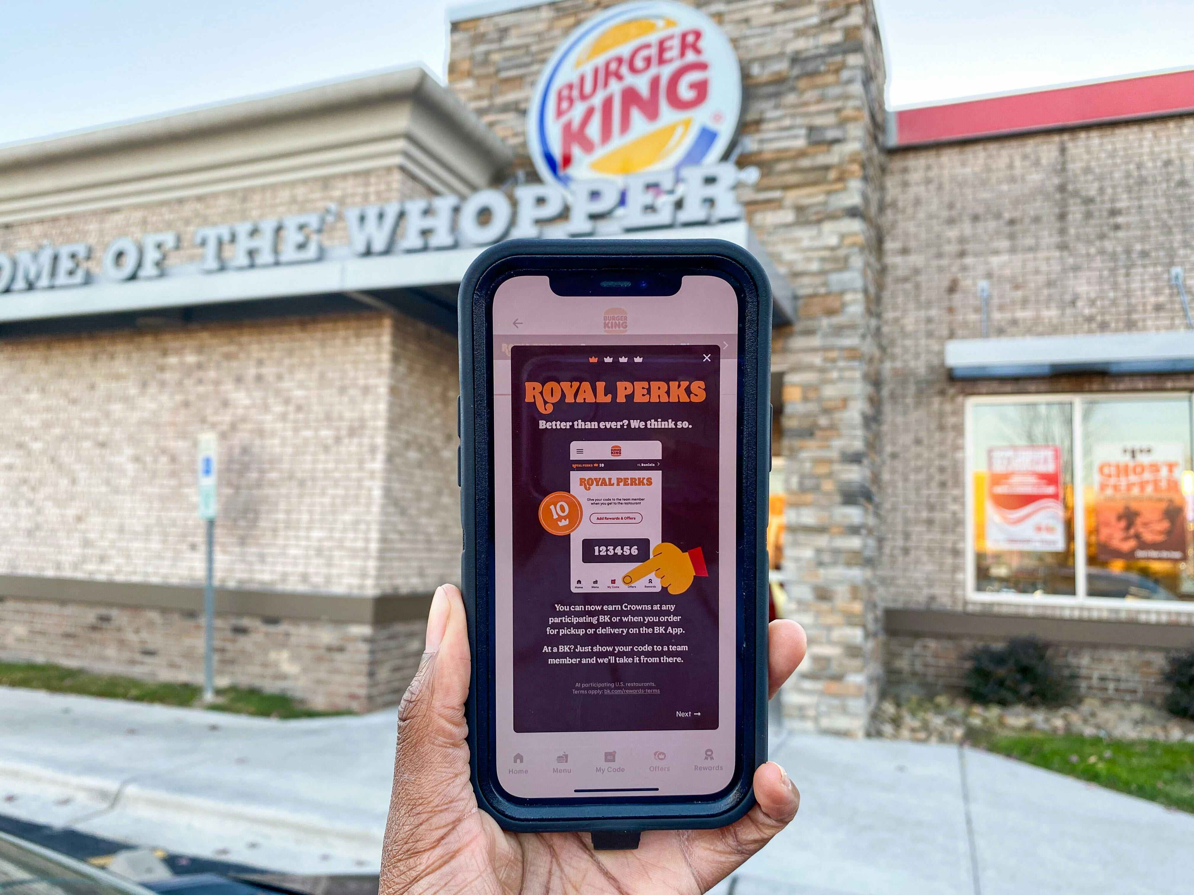 A cell phone displaying the Burger King Royal Perks app being held up in front of a Burger King restaurant storefront.