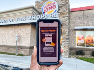 A cell phone displaying the Burger King Royal Perks app being held up in front of a Burger King restaurant storefront.