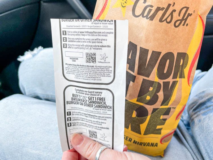 a hand holding a carls jr receipt next to a to go bag on lap