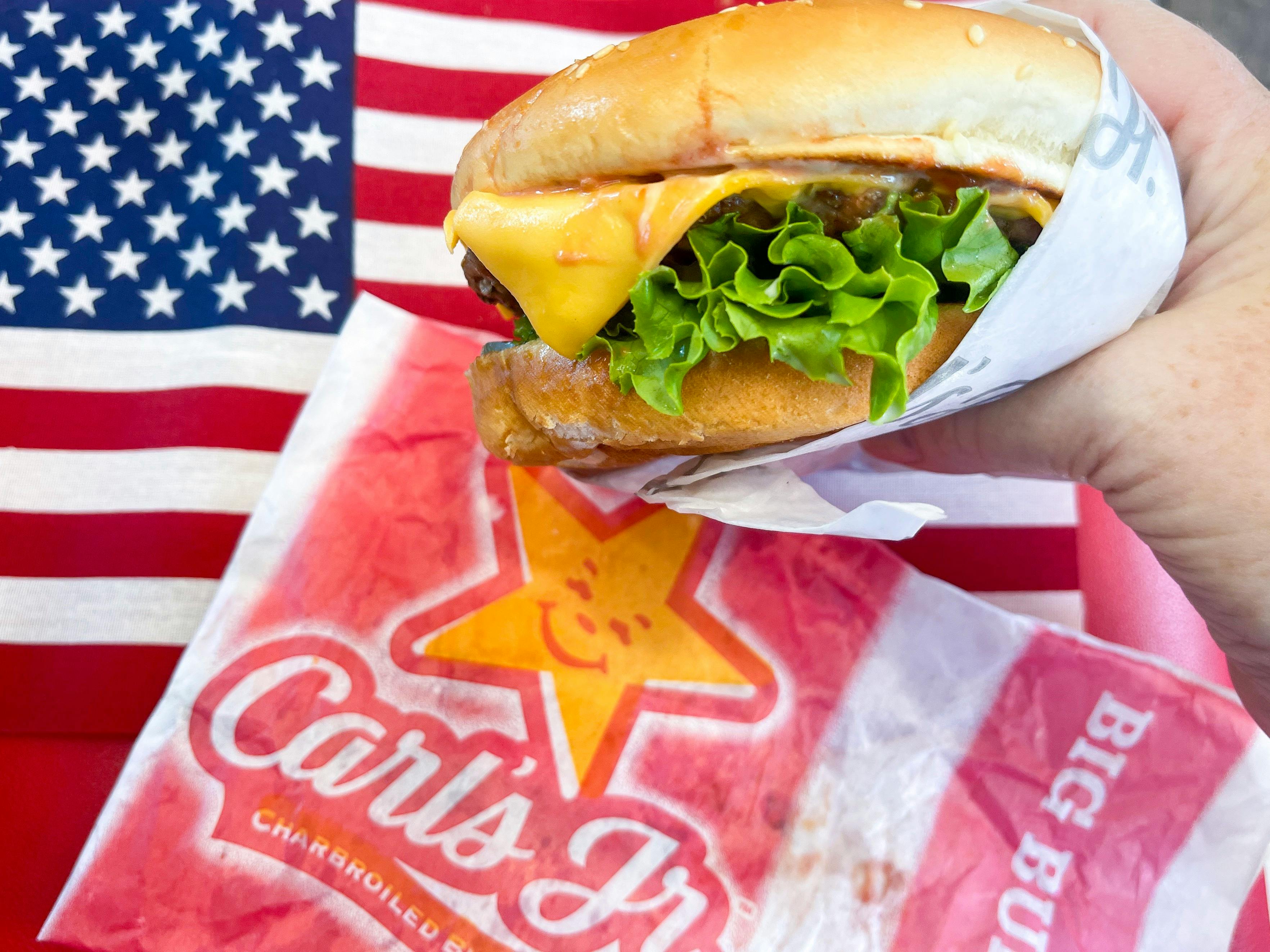 a hand holding a cheese burger in front of an american flag with a carls jr wrapper beside
