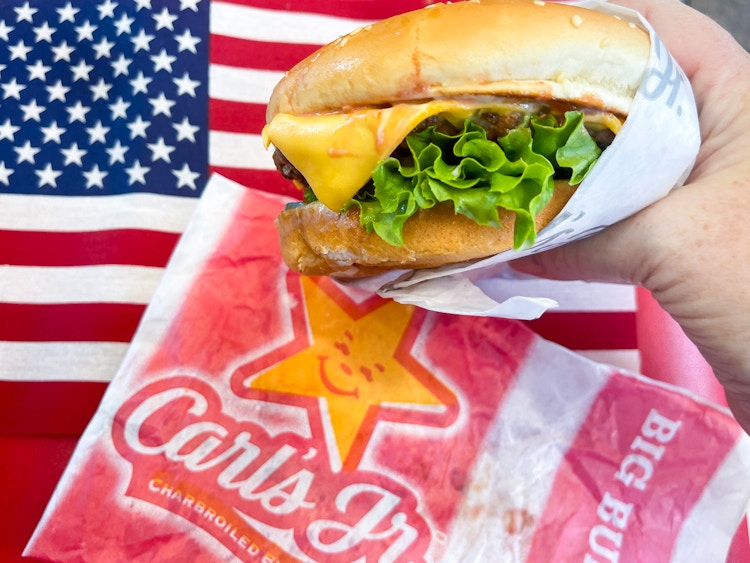a hand holding a cheese burger in front of an american flag with a carls jr wrapper beside